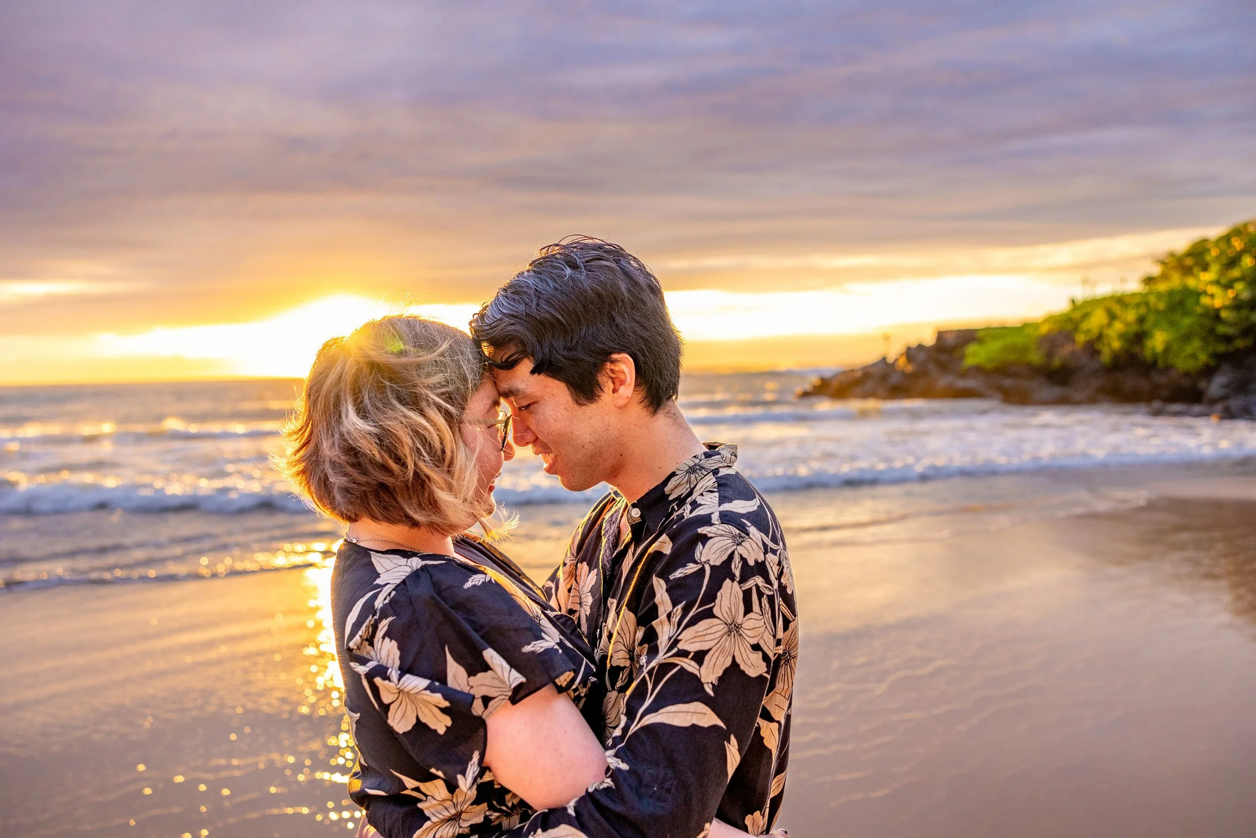 A couple embracing on a beach at sunset, with the sun setting over the ocean and rocky cliffs in the background.