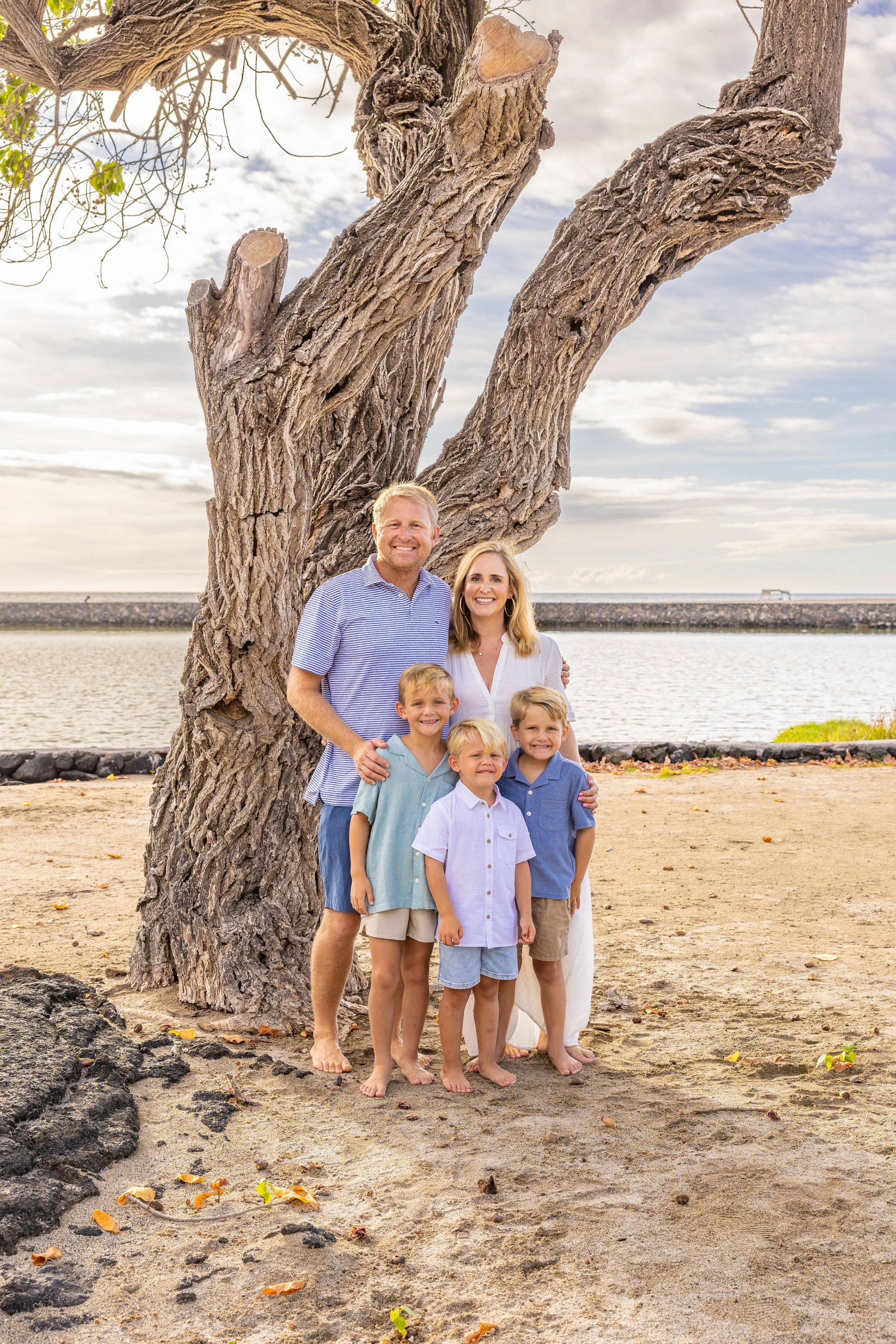 A smiling family of six, including two adults and four children, standing on a sandy beach in front of a large, bare tree with a water body and cloudy sky in the background.
