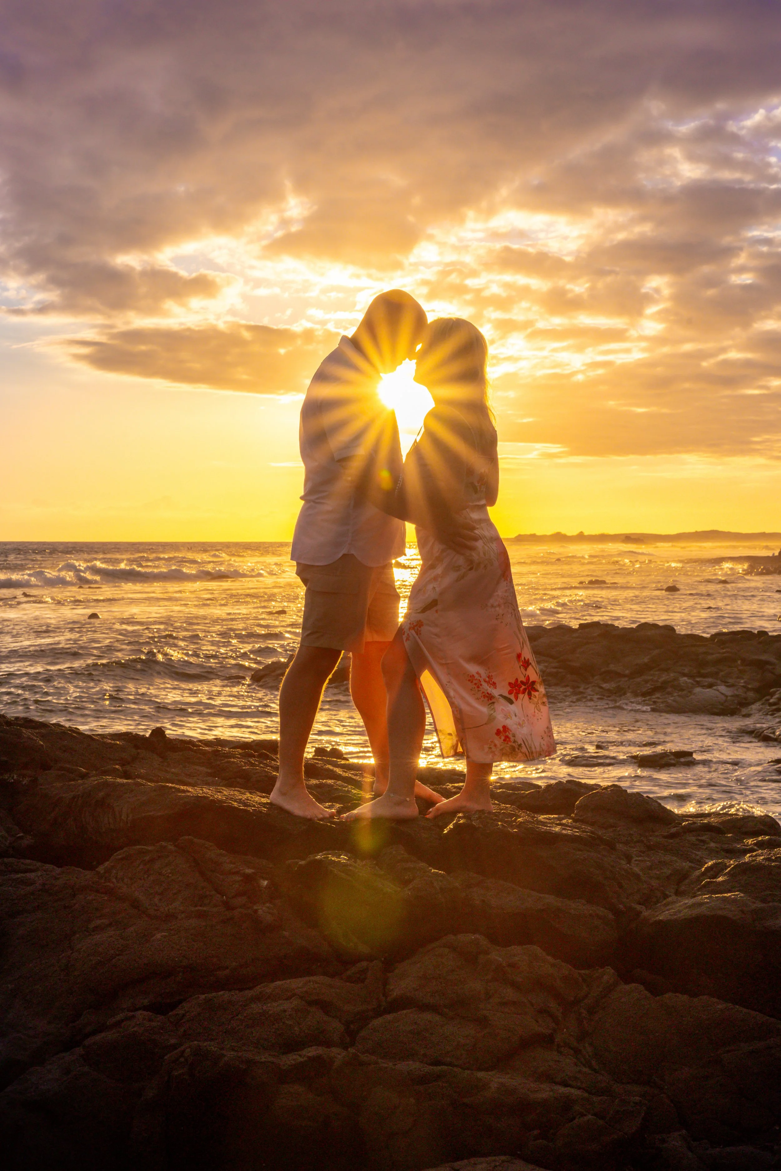 A couple standing on rocks by the ocean at sunset, with their foreheads touching and facing each other, the sun shining between them.