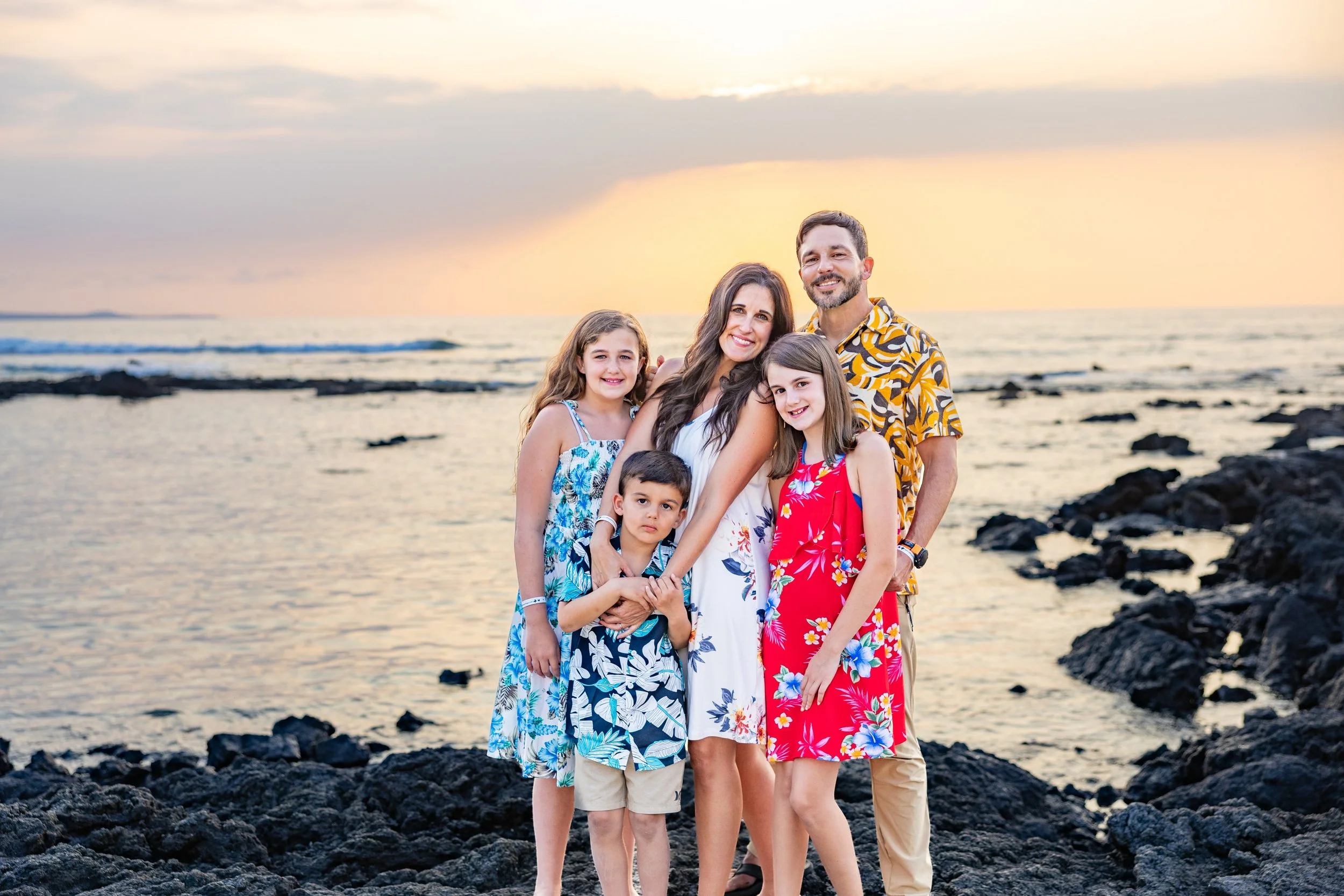 Family of six standing on rocks by the ocean at sunset, smiling and posing for the photo