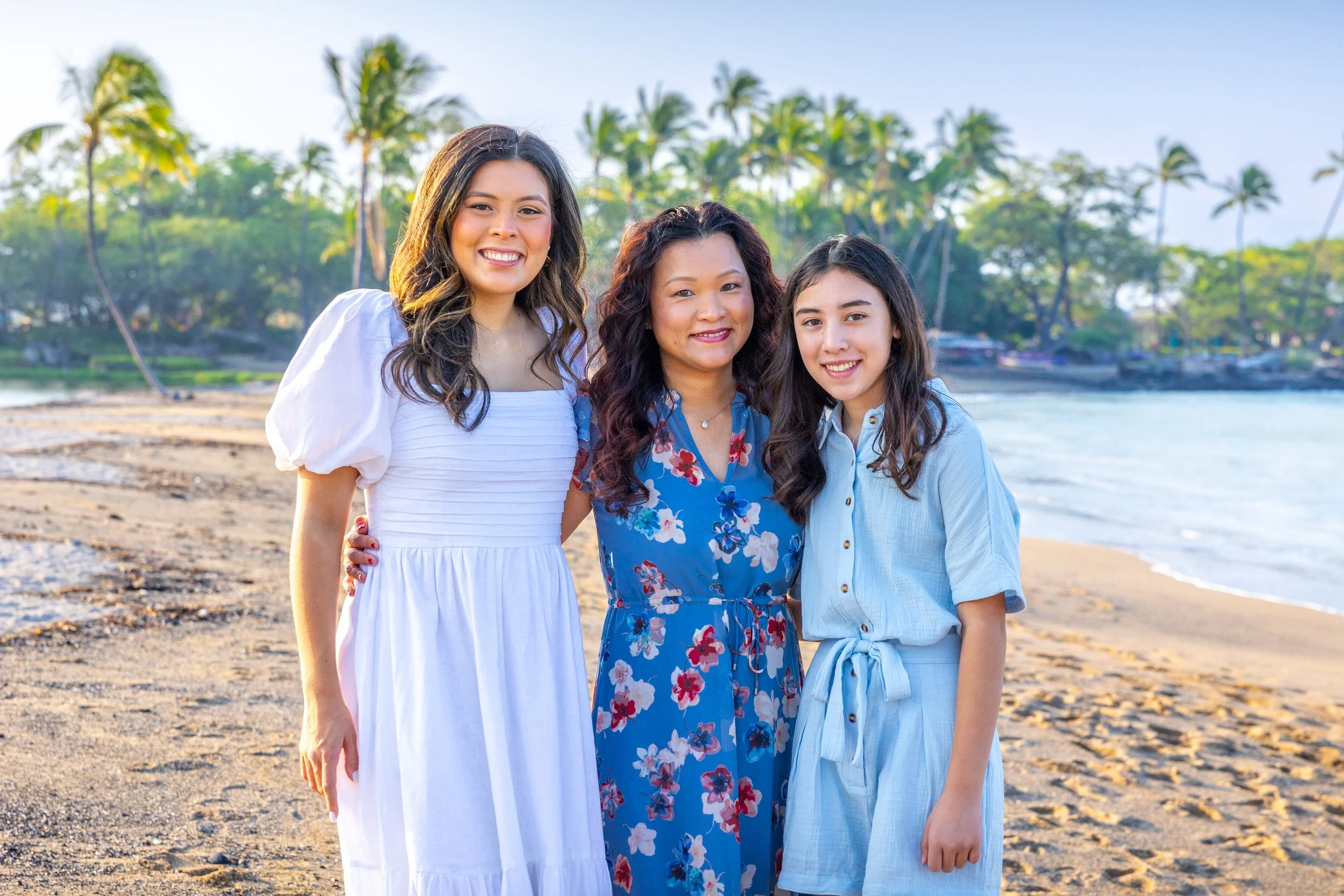Three women standing close together on a beach with palm trees in the background, smiling at the camera.
