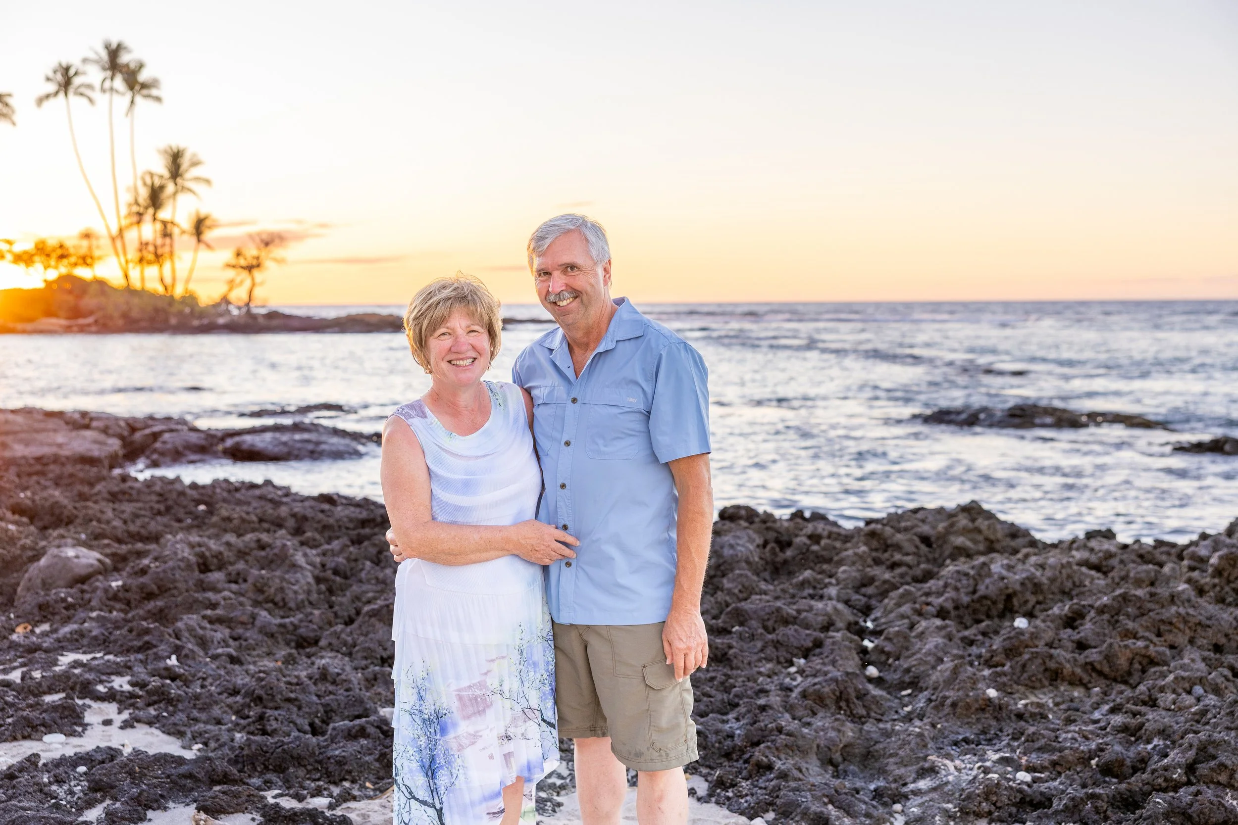A smiling elderly couple standing on rocky beach during sunset with ocean waves in background.