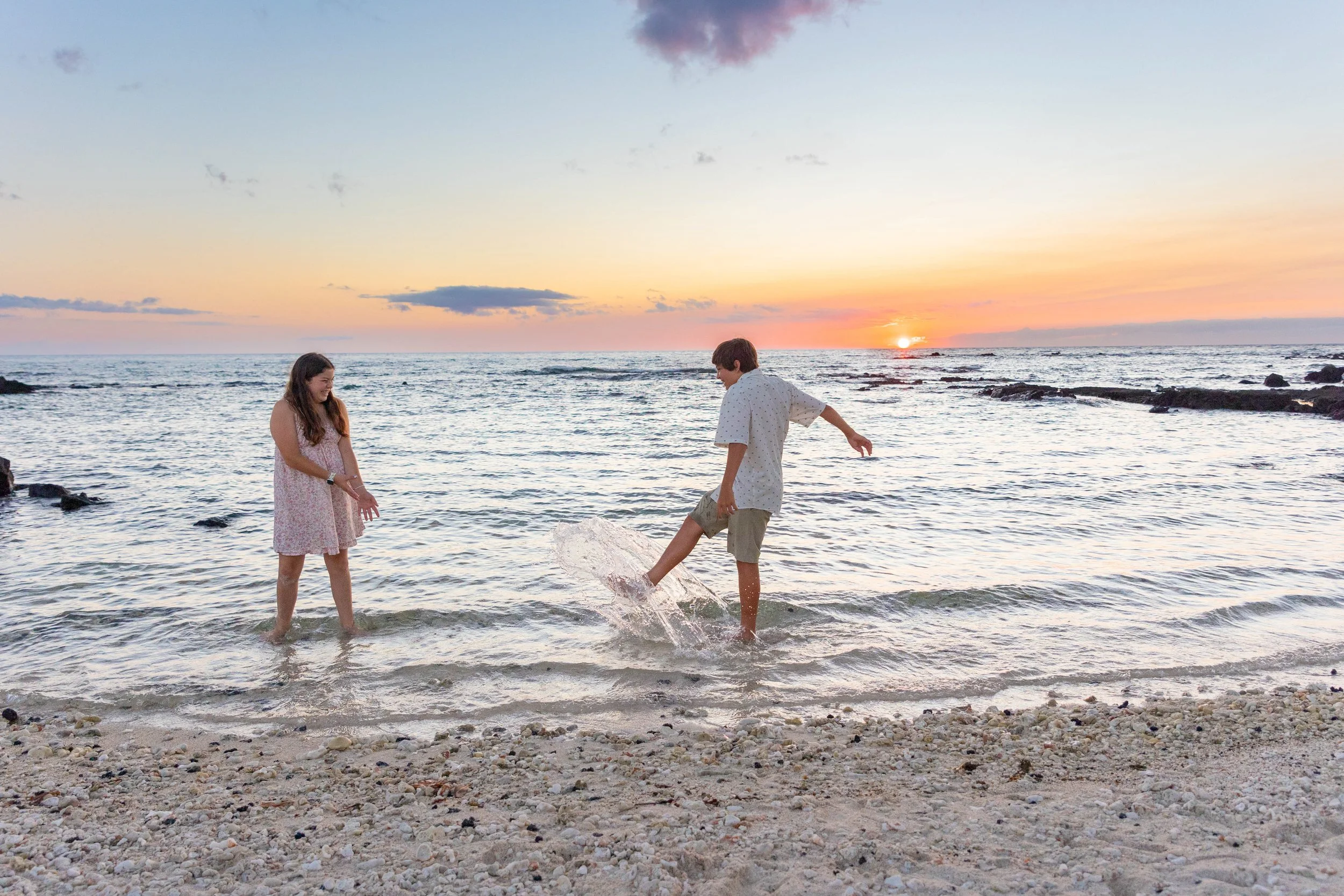 A boy and girl playing in shallow ocean water at sunset on a rocky beach, with the sun setting on the horizon.