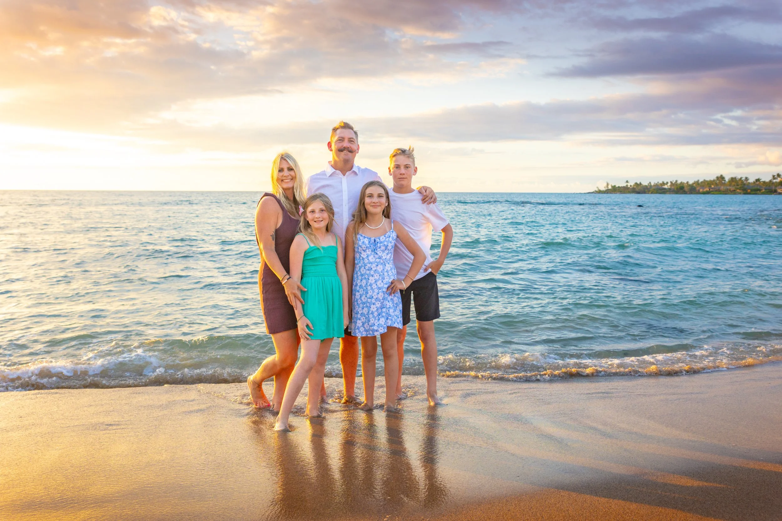 Family of five standing on a beach at sunset, with the ocean in the background, smiling and posing for the photo.