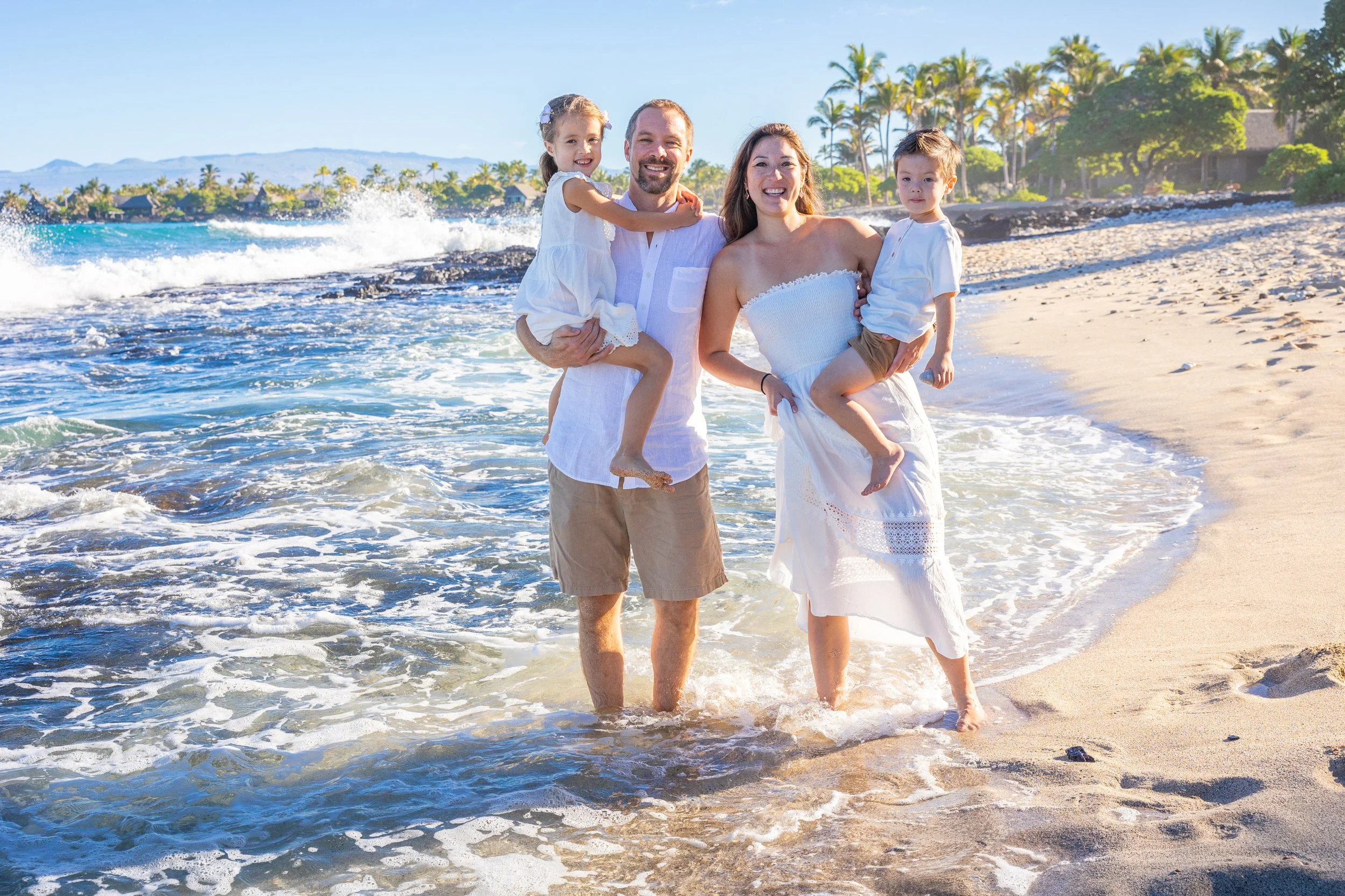 A happy family of four enjoying a day at the beach, with ocean waves, sand, and palm trees in the background.