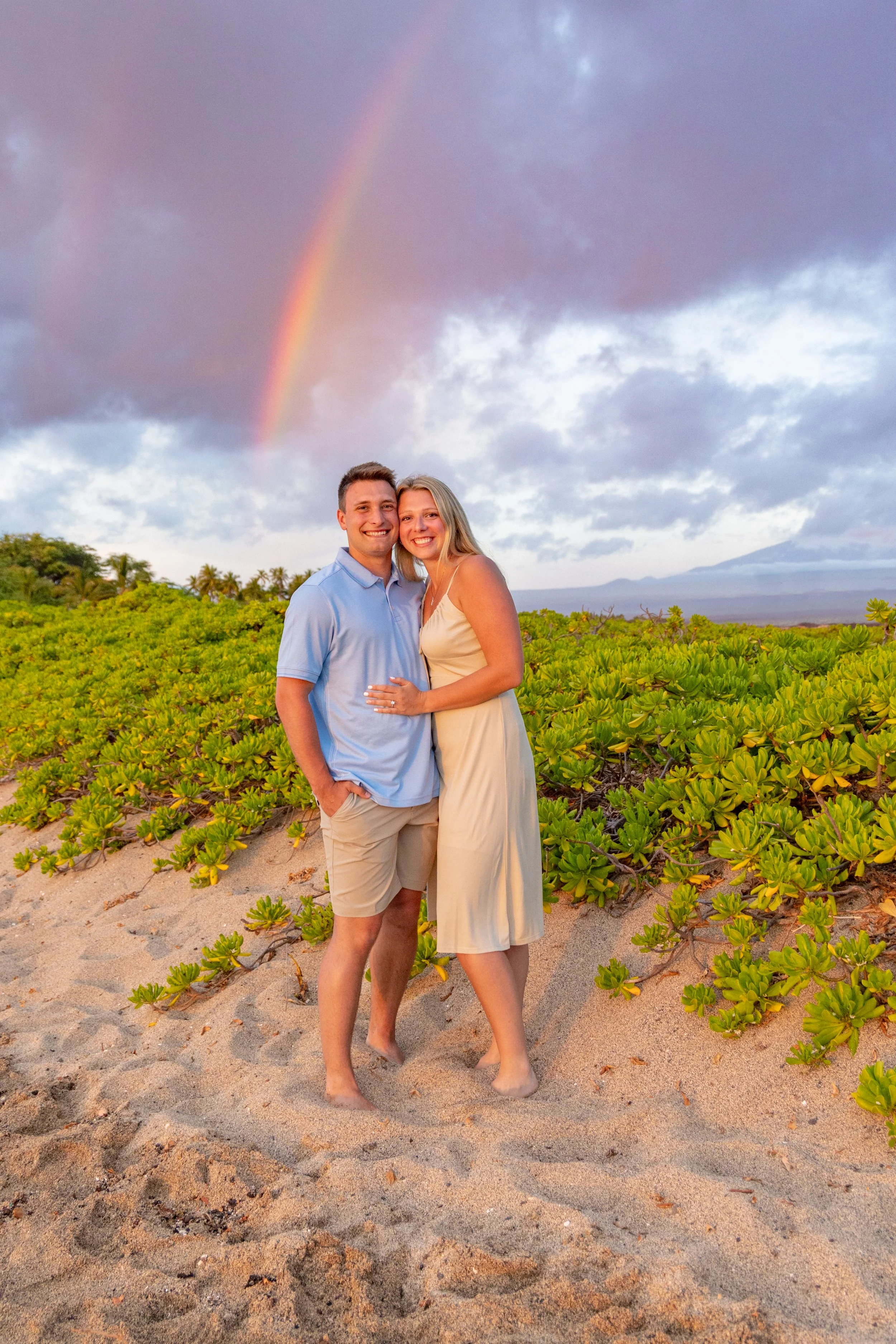 A happy couple standing on a sandy beach with green bushes in the background, under a cloudy sky with a rainbow.