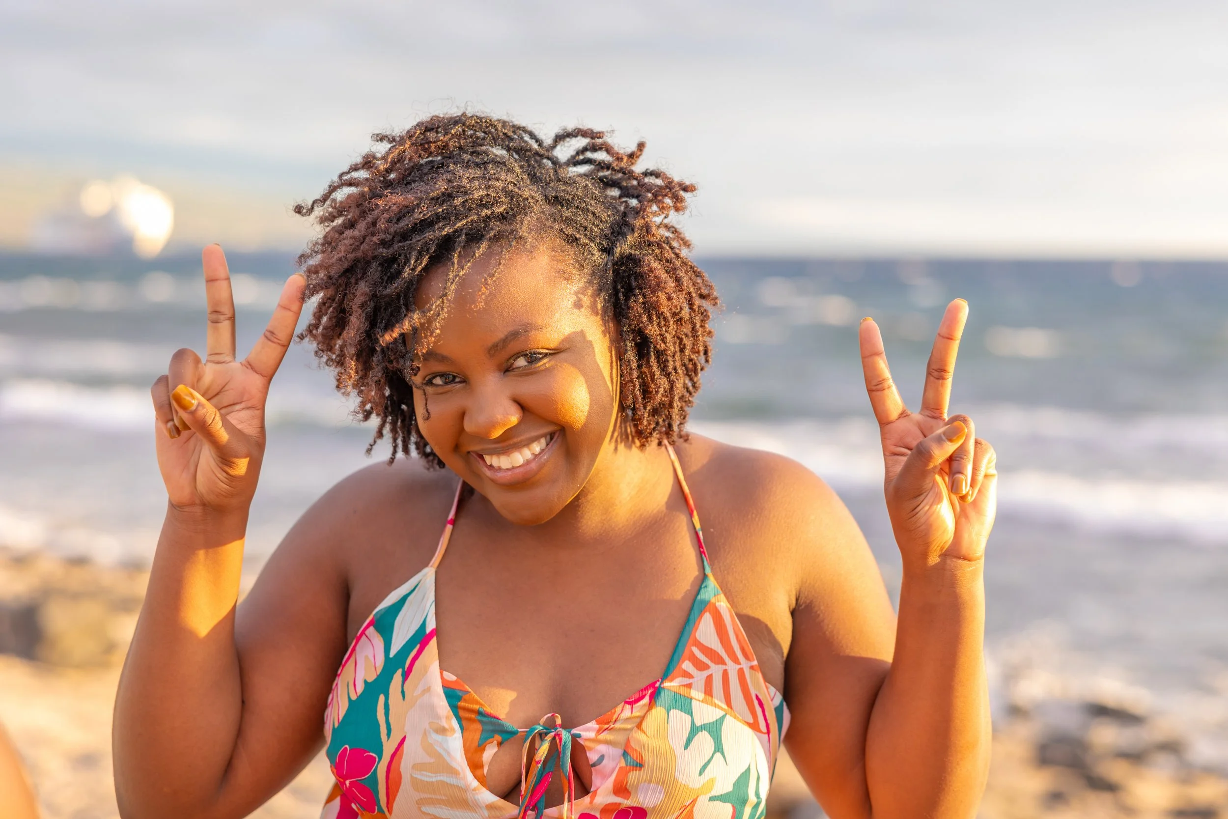 A smiling woman with curly hair making peace signs with both hands at the beach, wearing a colorful swimsuit.