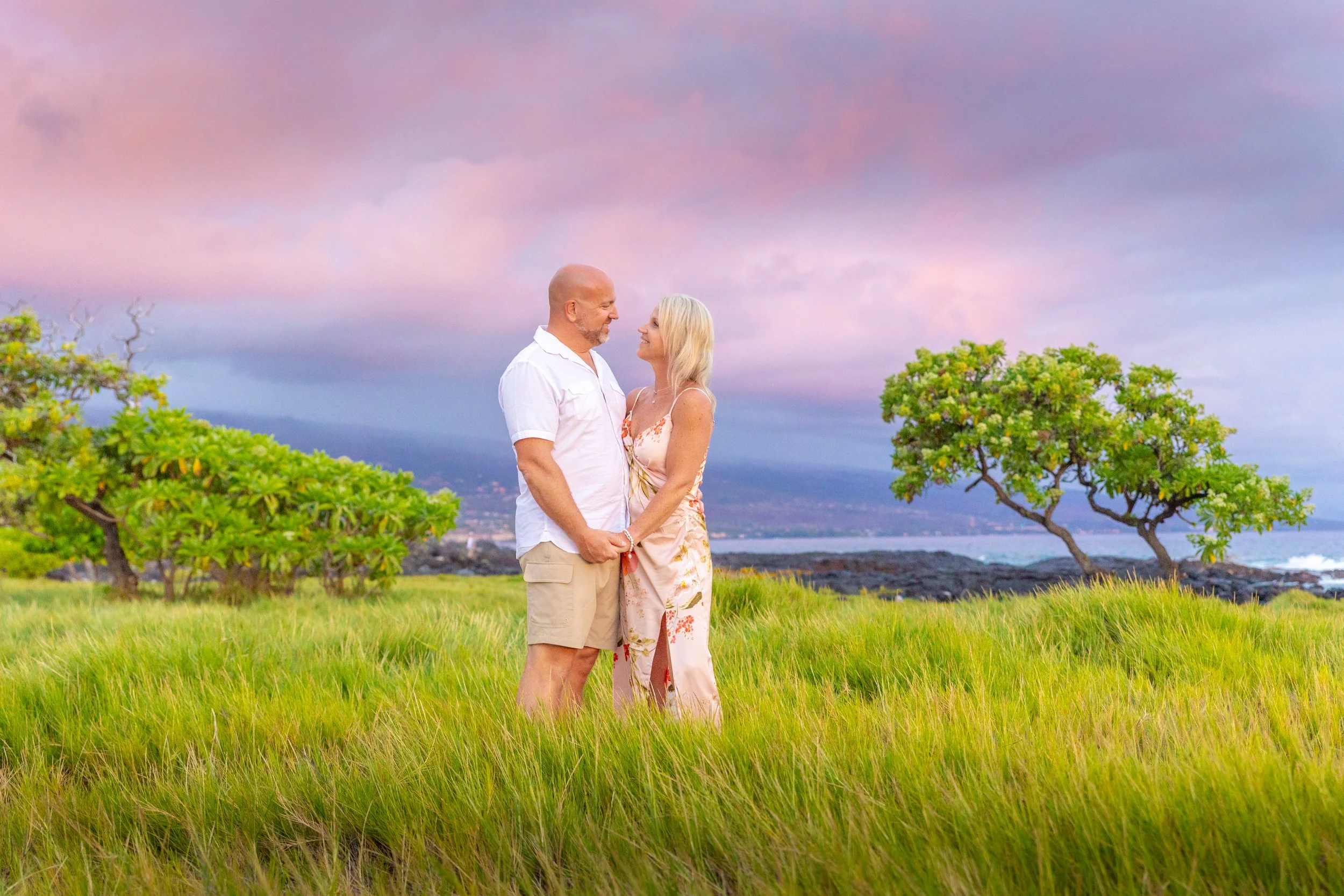 A couple standing in a grassy field holding hands and gazing into each other's eyes, with trees and a rocky coastline in the background and a pinkish-purple sky during sunset.