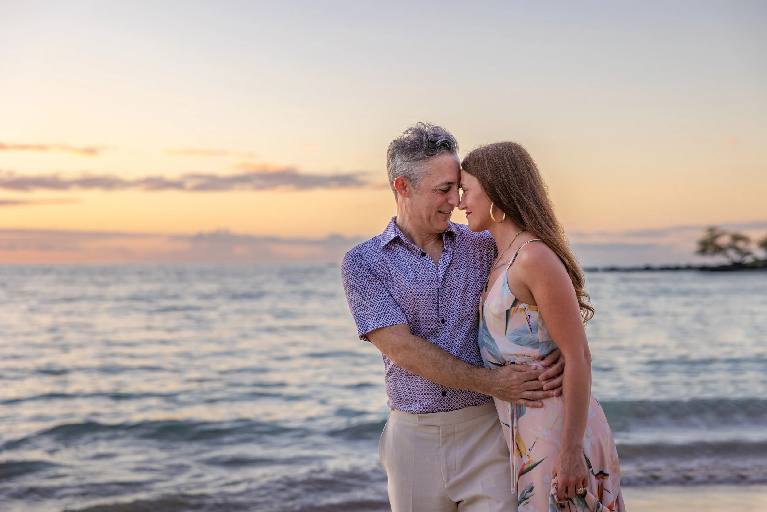 A middle-aged man and a woman embrace on a beach at sunset, their foreheads touching, with water and a colorful sky in the background.