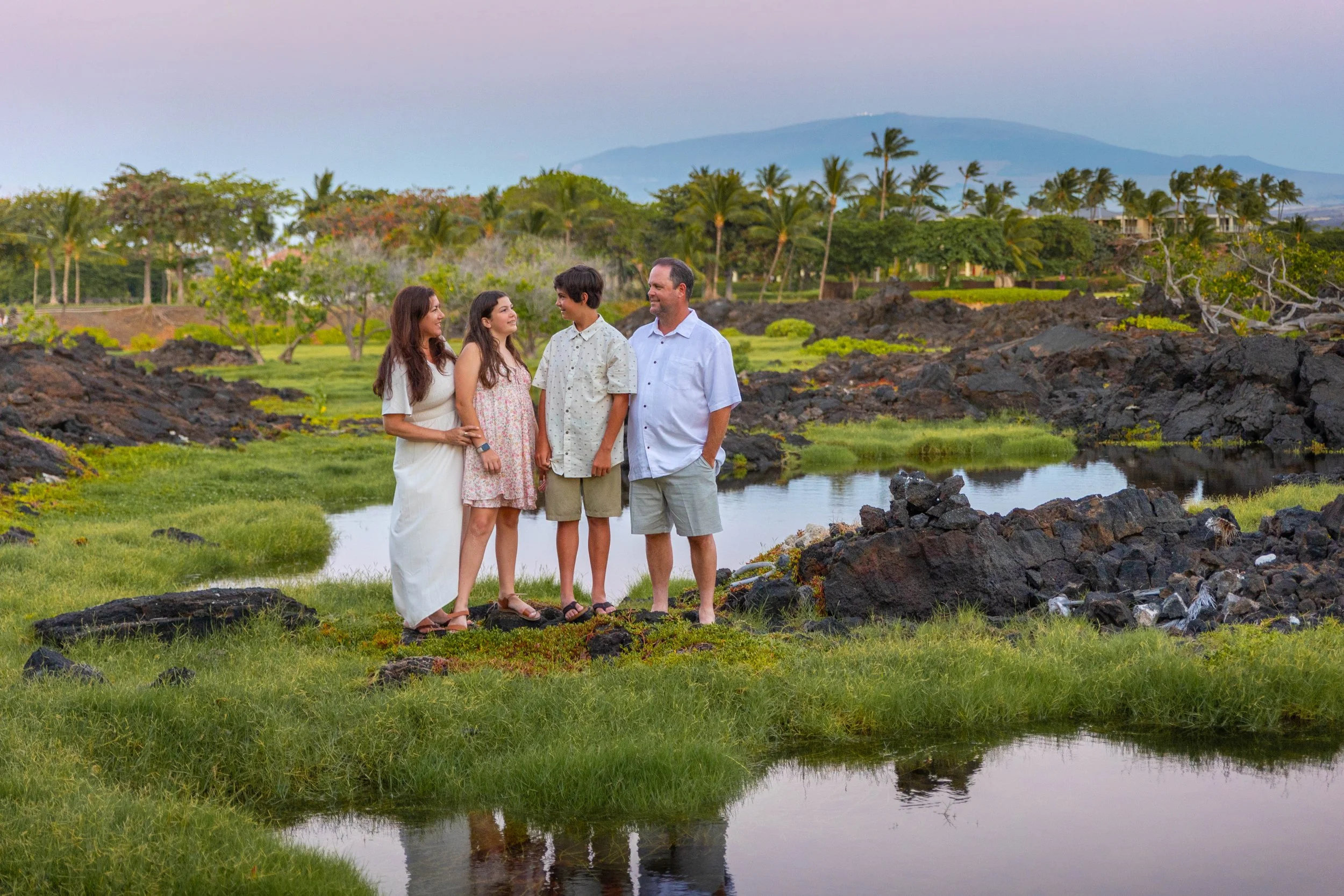 Family of five standing on grass near a small body of water in a scenic, tropical landscape with palm trees and mountains in the background.