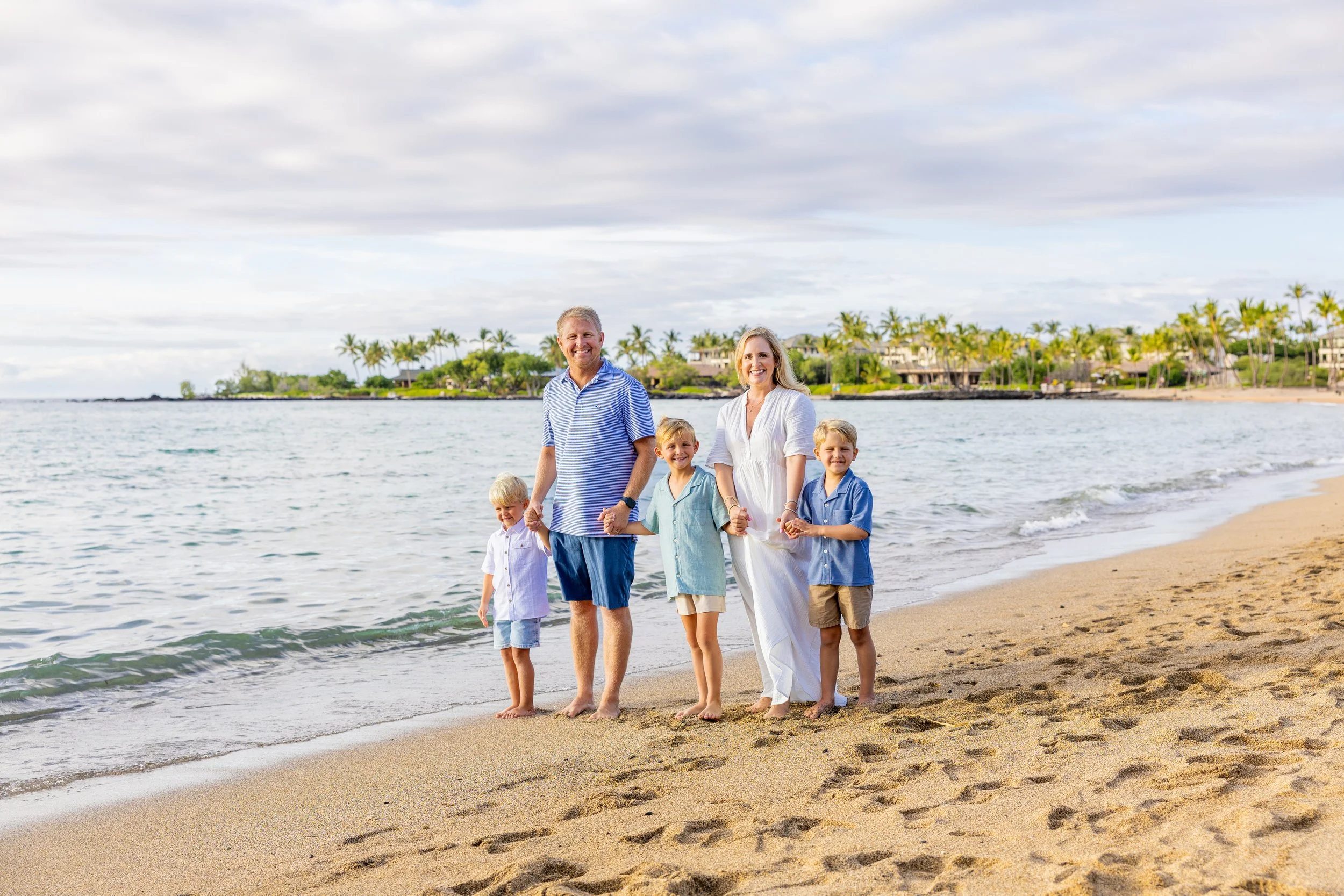 A happy family of six standing on the beach holding hands, with waves and palm trees in the background.