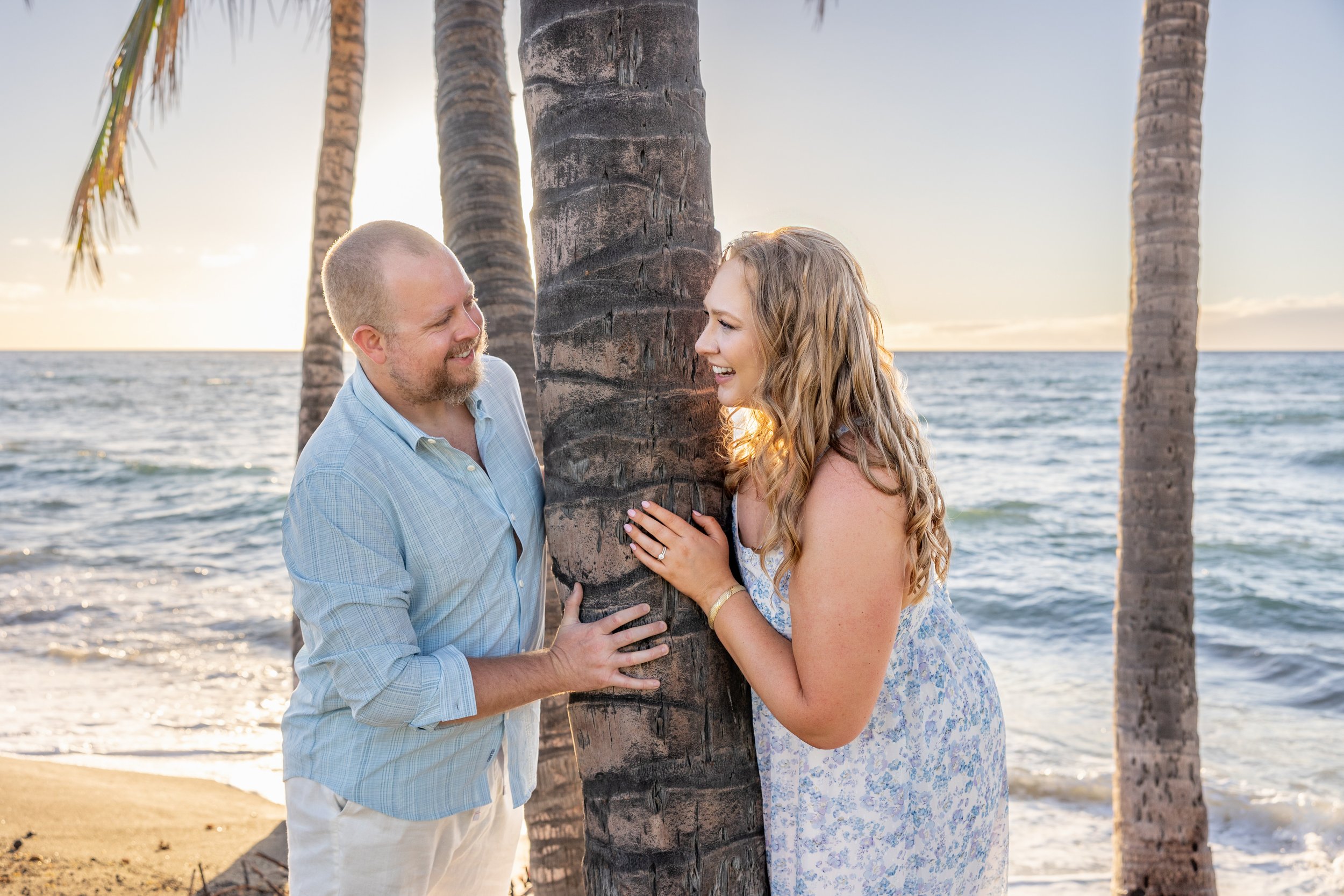 A couple stands on the beach near palm trees, smiling and touching a tree trunk as the sun sets over the ocean.