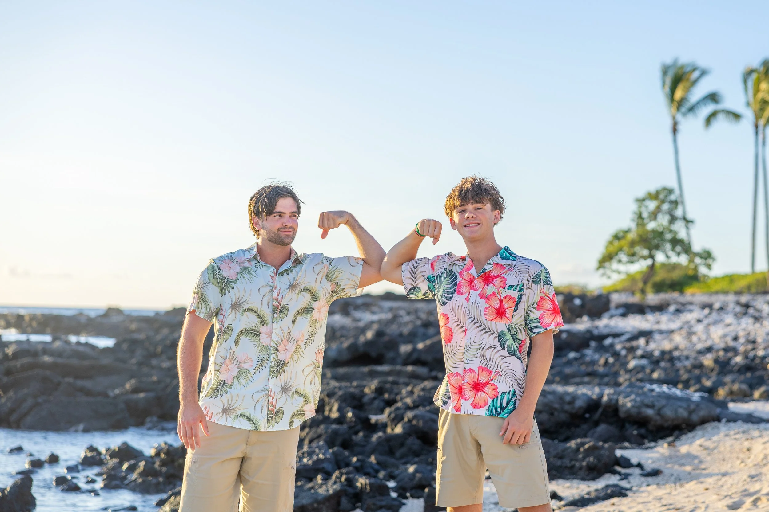 Two young men in Hawaiian shirts and shorts posing with flexed arms on a beach with rocks and palm trees in the background.