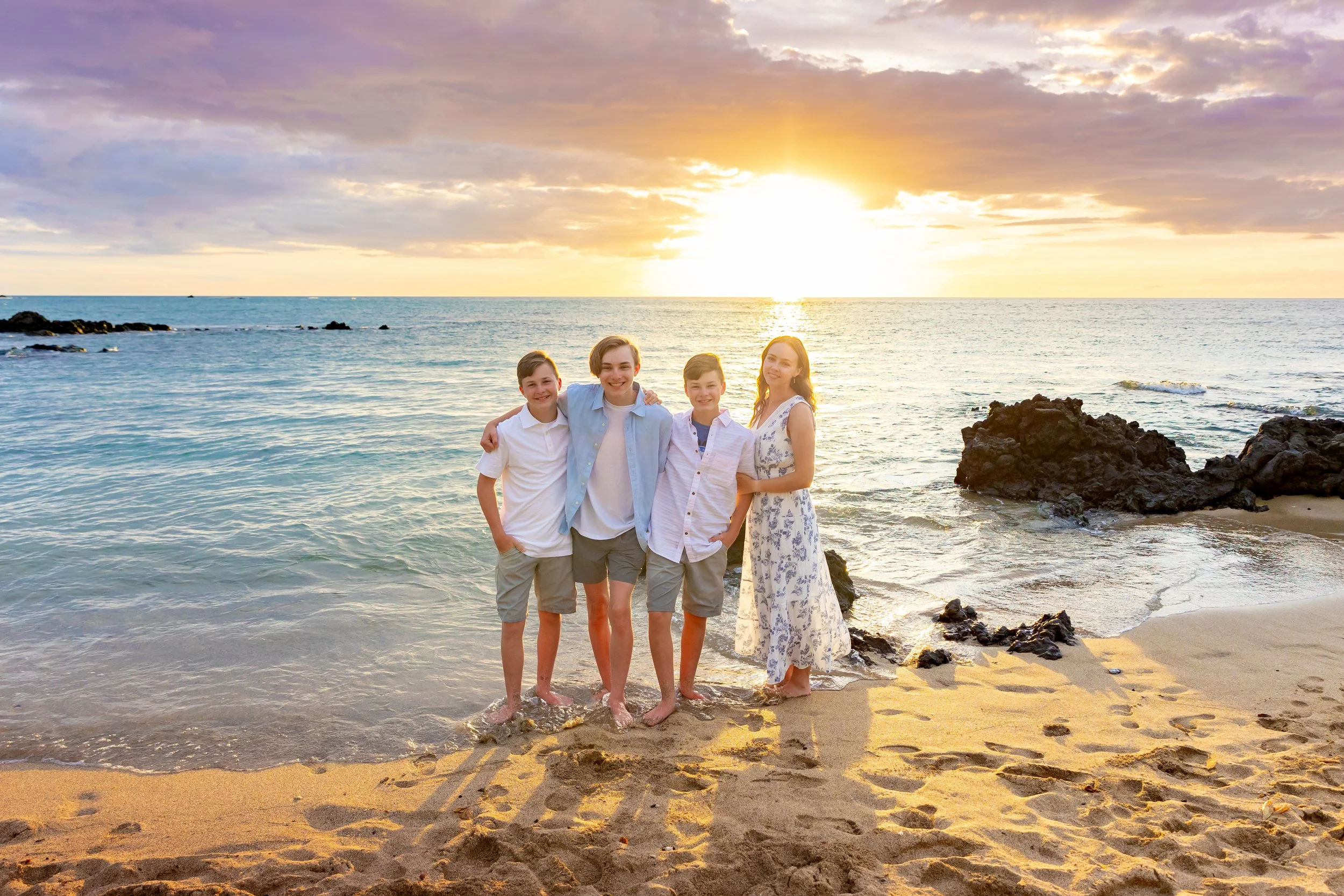 A family of four standing on the beach during sunset, with the ocean and rocks in the background.