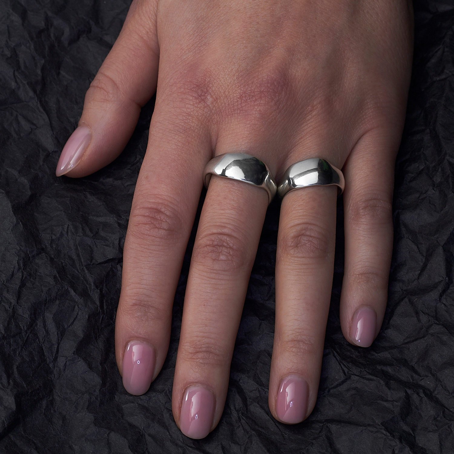 A female hand with light pink nail polish wears two thick silver rings against a textured black background.