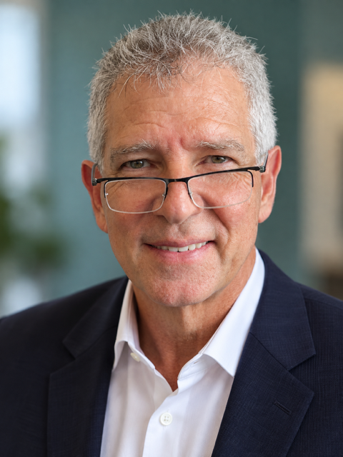 A middle-aged man with gray hair wearing a navy suit, white shirt, and a patterned tie, smiling at the camera.