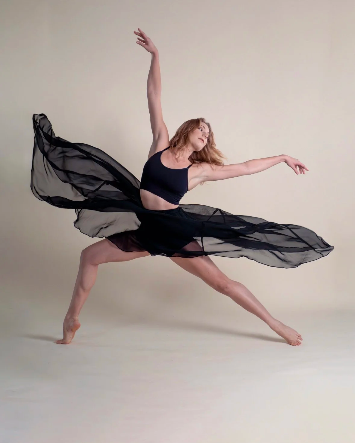 A female dancer in a black sports bra and flowing black skirt posing dynamically against a plain background.