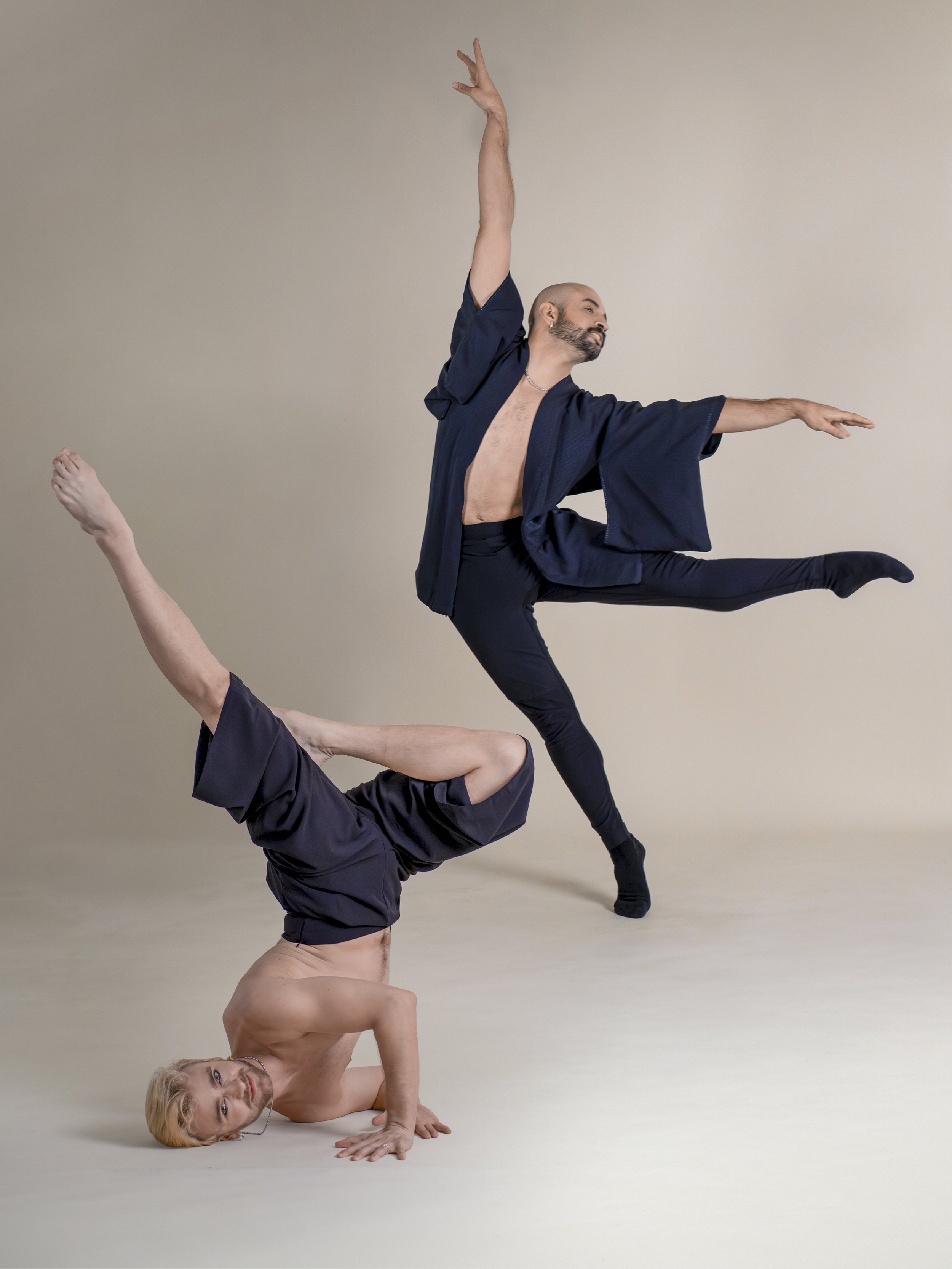 Two men, one shirtless with blond hair doing a one-handed headstand, and the other in a navy robe and black pants dancing in a studio with a plain beige background.