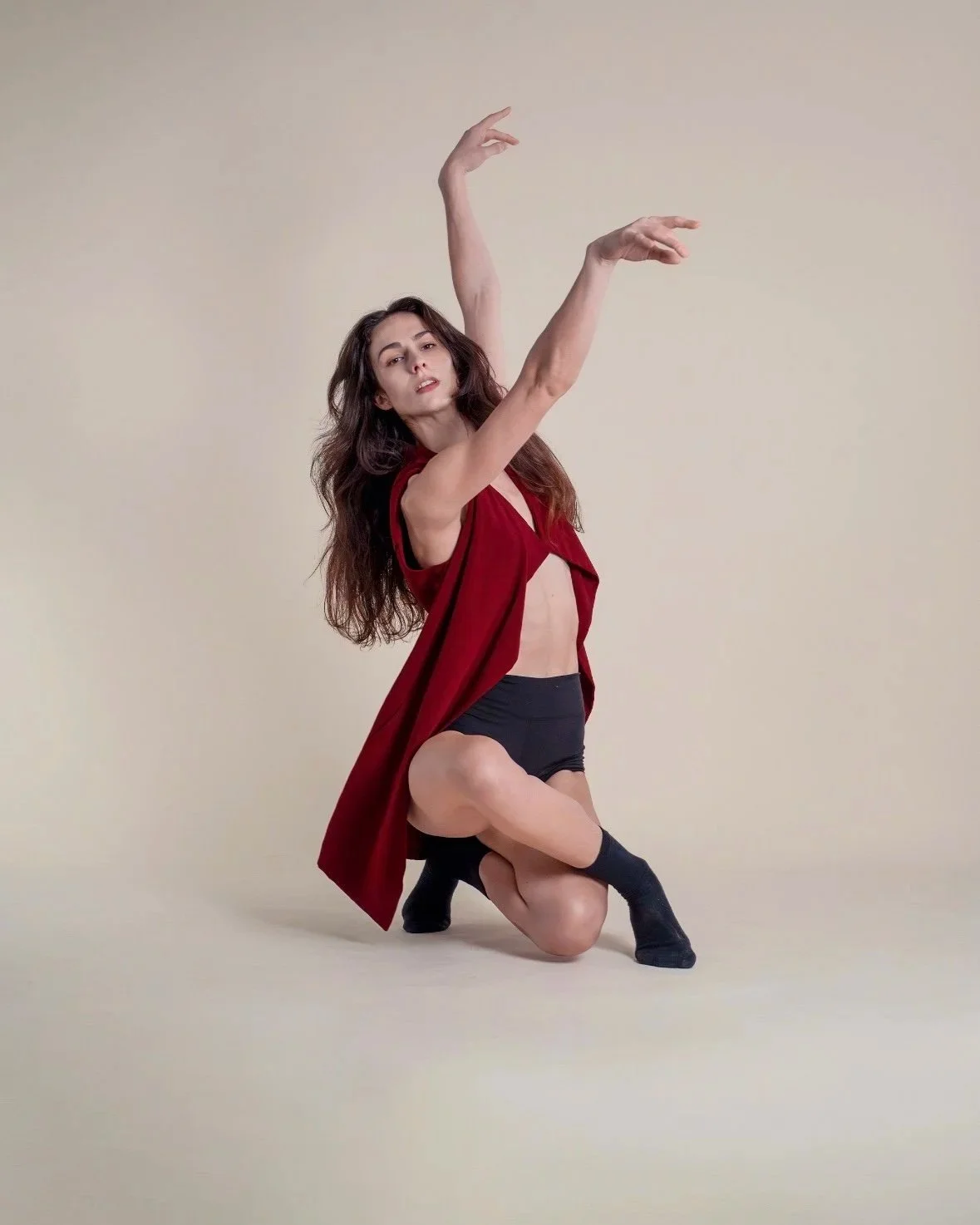 A woman with long dark hair is posing on a plain beige backdrop, wearing a red sleeveless open vest, black shorts, and black socks, kneeling with one knee on the ground and her arms raised in a dance pose.