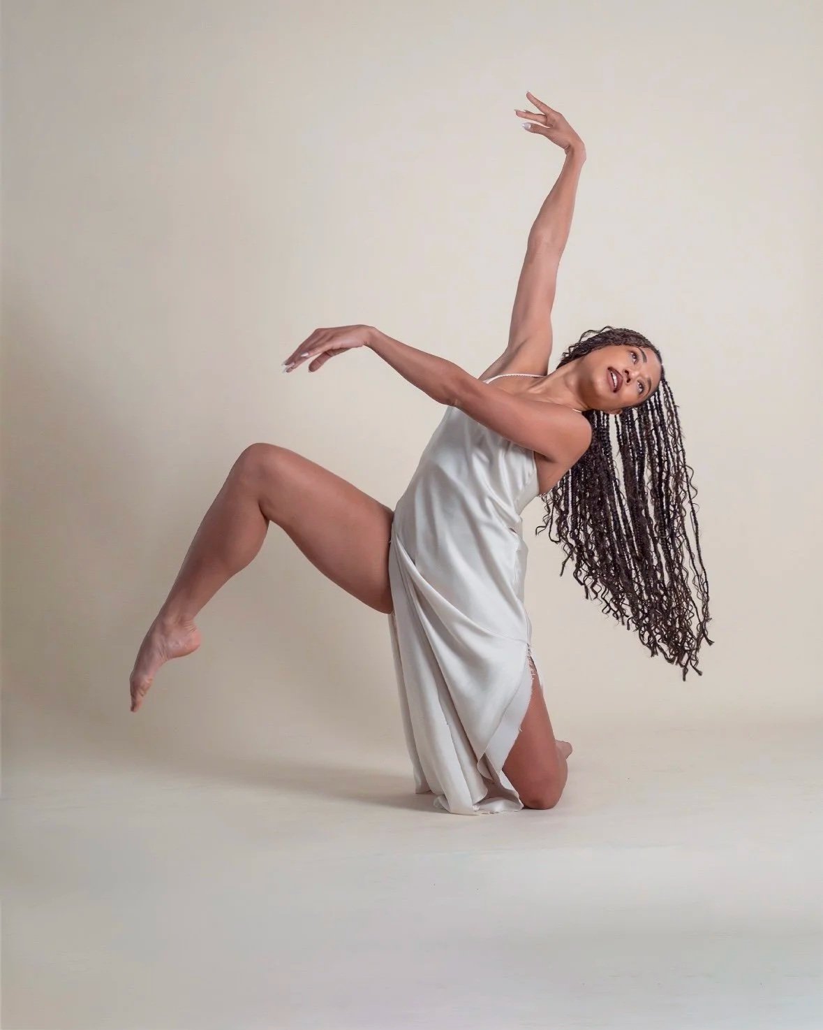 A dancer with long, curly hair poses gracefully on a plain beige background, wearing a silky, white slip dress.