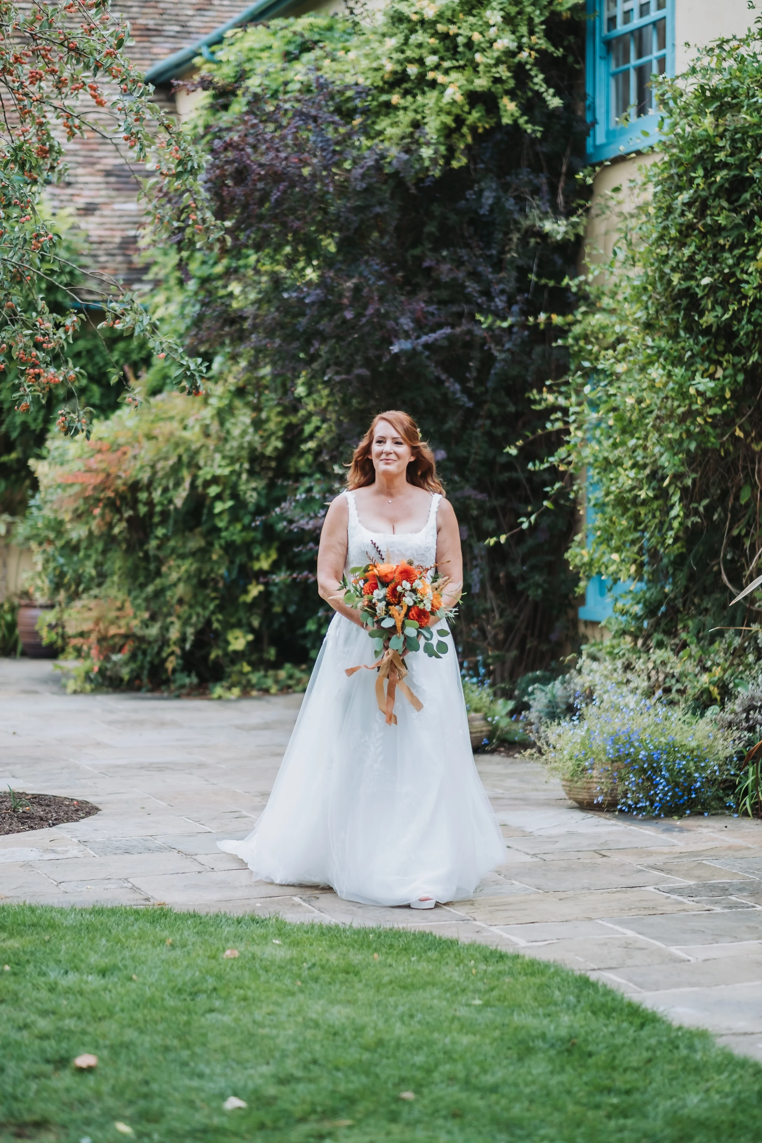 A woman in a white wedding dress holding a bouquet of orange and white flowers standing outdoors on a stone patio surrounded by lush green bushes and flowering plants.