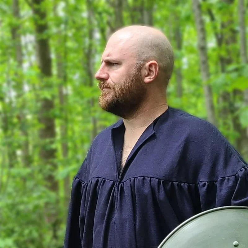 A chest-up photo of Zeb Mientkiewicz. He is wearing a navy blue ren faire shirt and holding a large metal shield. His head is clean shaven and he has a beard.