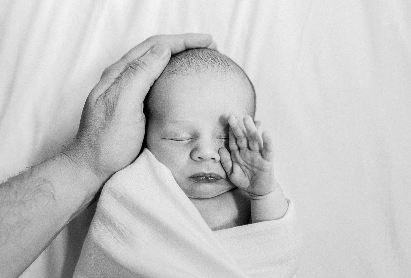 Baby boy wrapped in blanket and rubbing his eyes, while dad's hand holds his head by Chicago Baby Photographer