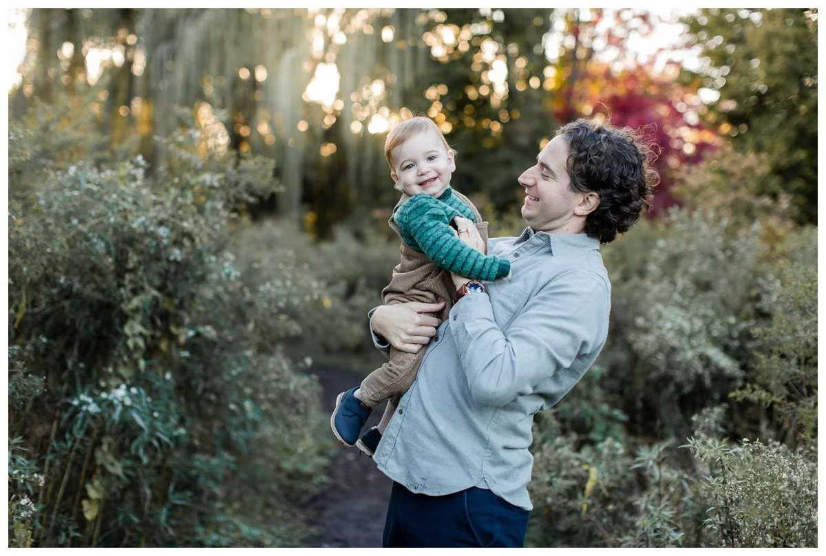 Father holding baby boy while giggles with a willow in the background and sun setting behind it