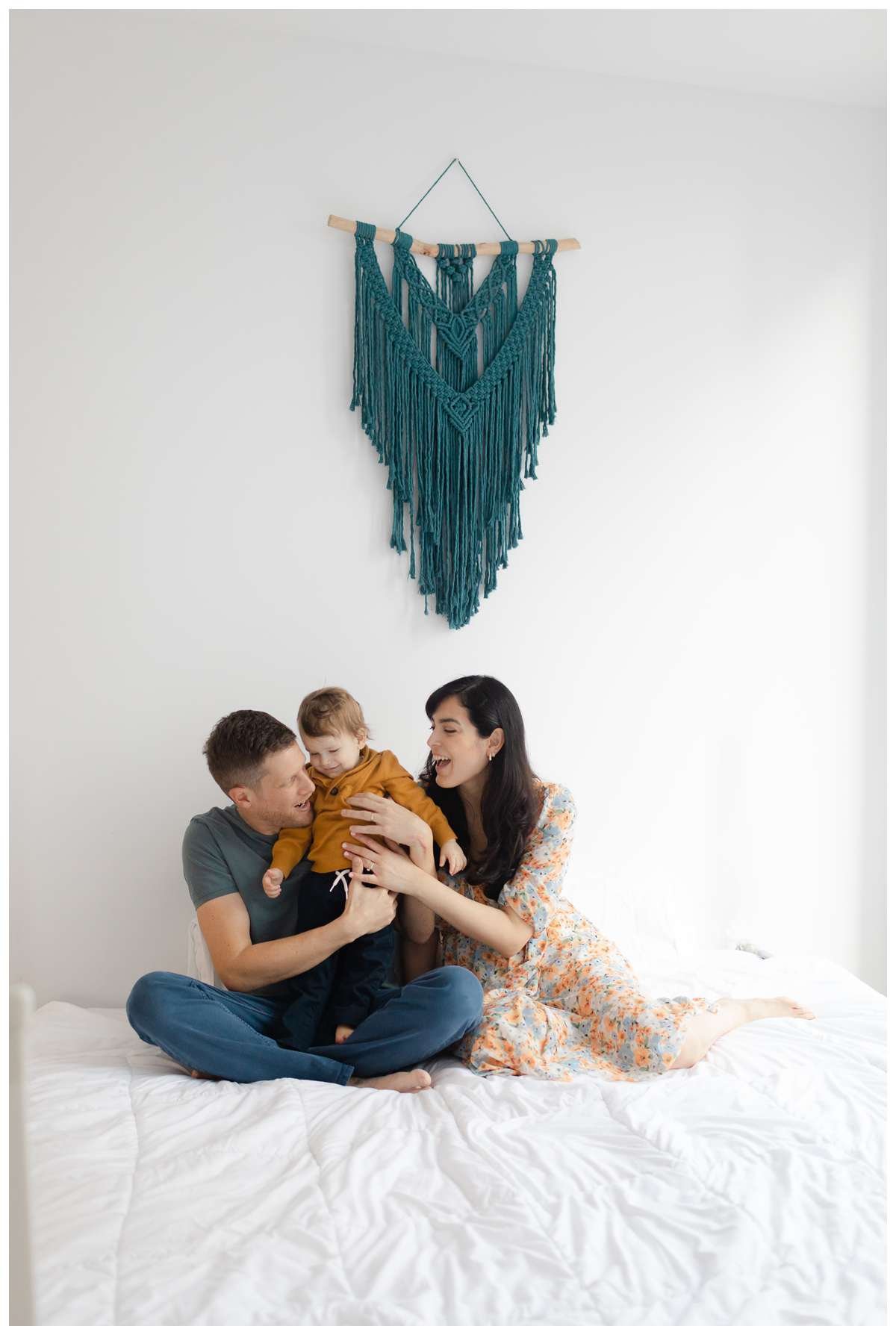 Parents playing with toddler boy on bed with a blue tapestry on the wall above