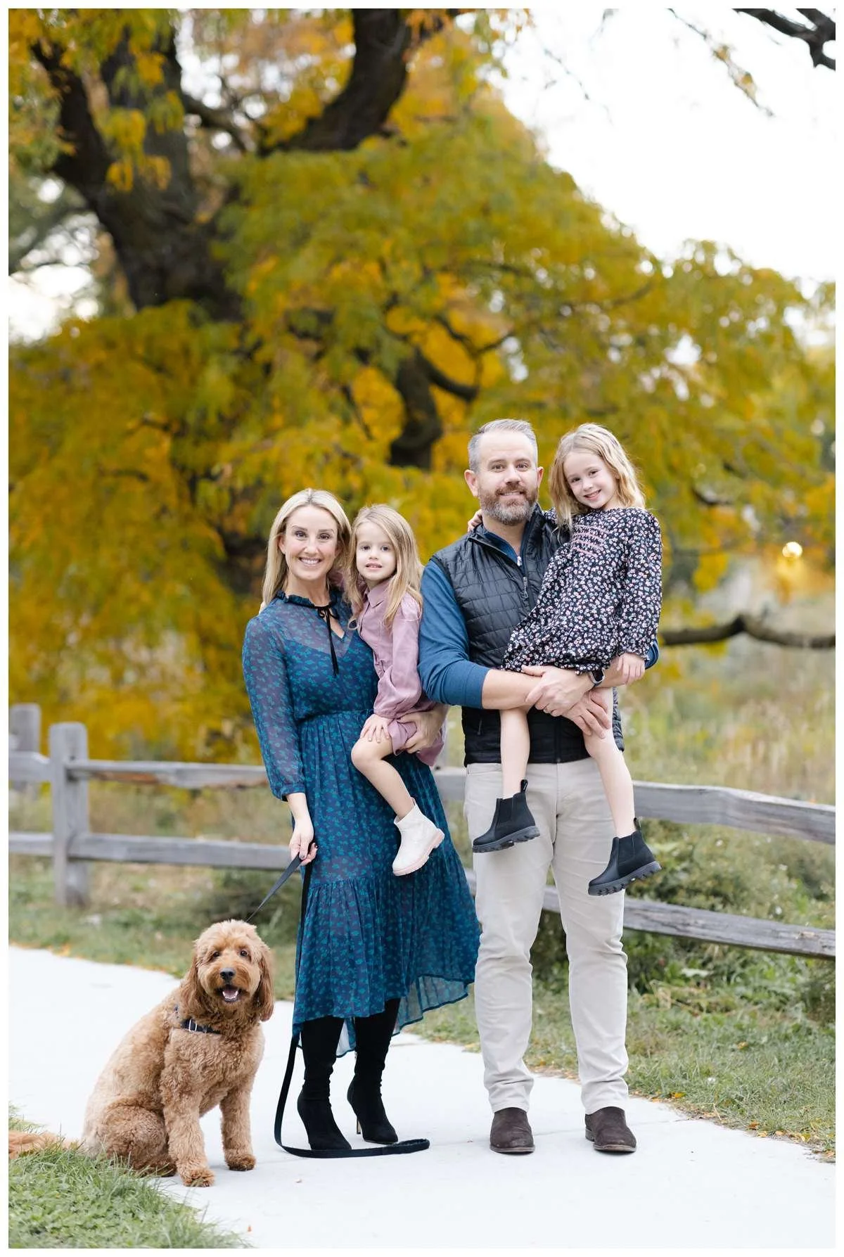 Parents carrying two young daughters and holding brown dog on leash against colorful foliage for Fall Family Photography Session