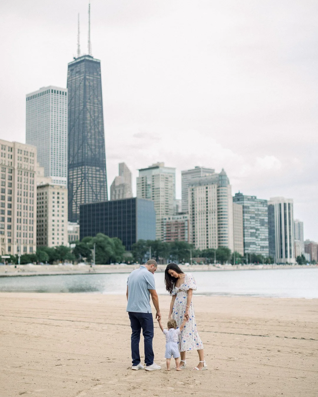 Parents holding toddler's hand as he walks on Ohio Street Beach with Chicago Skyline in background