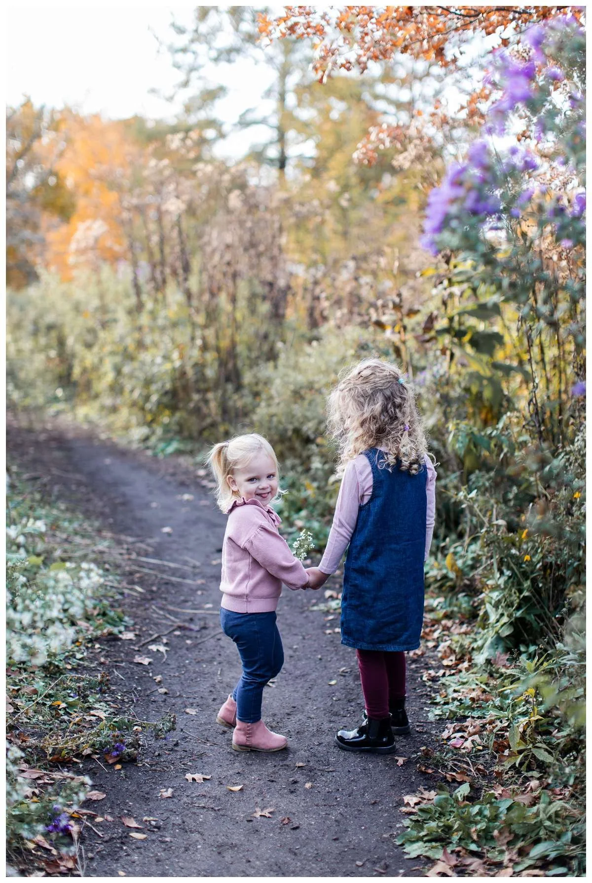 Two sisters holding hands along prairie path walk full of purple and yellow flowers
