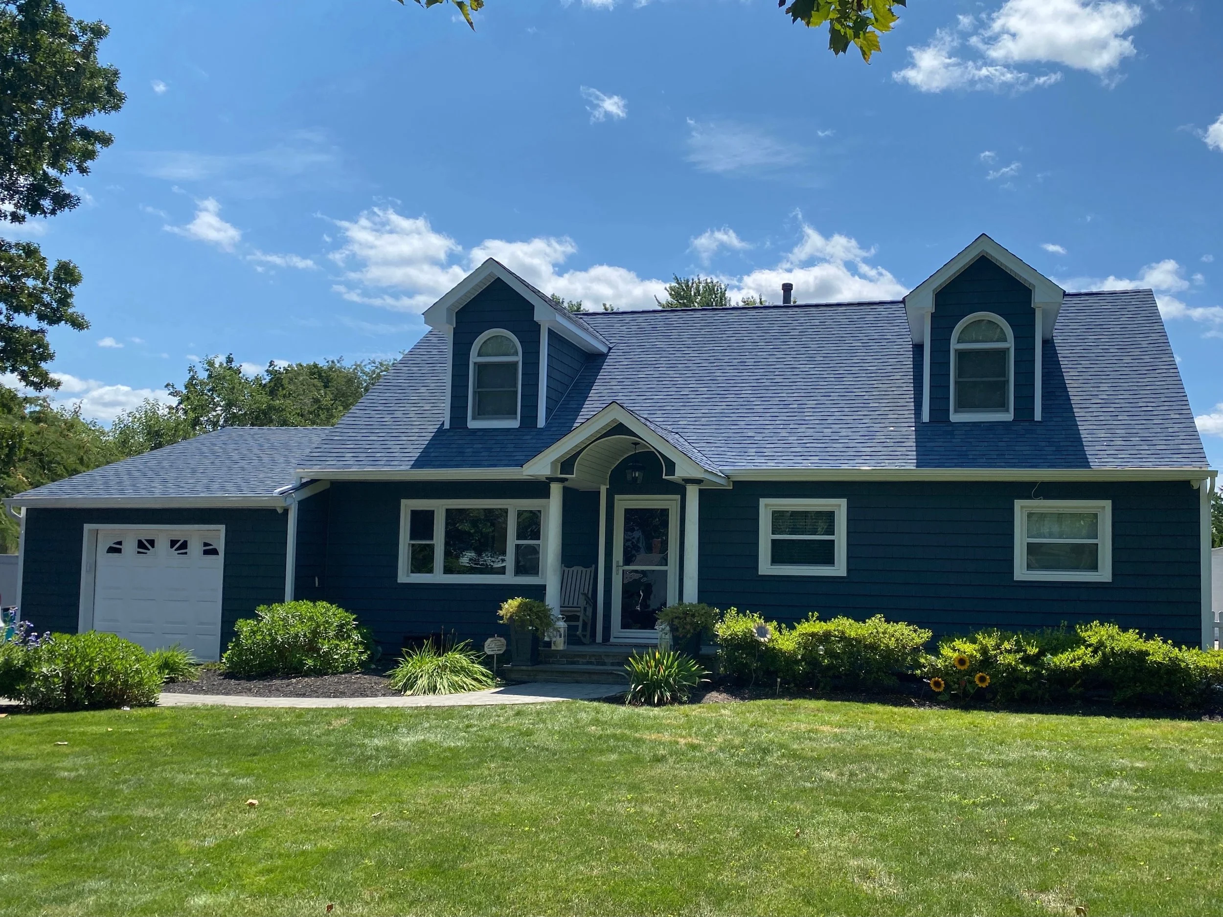 A blue house with a front porch, surrounded by a green lawn and garden, under a blue sky with some clouds.