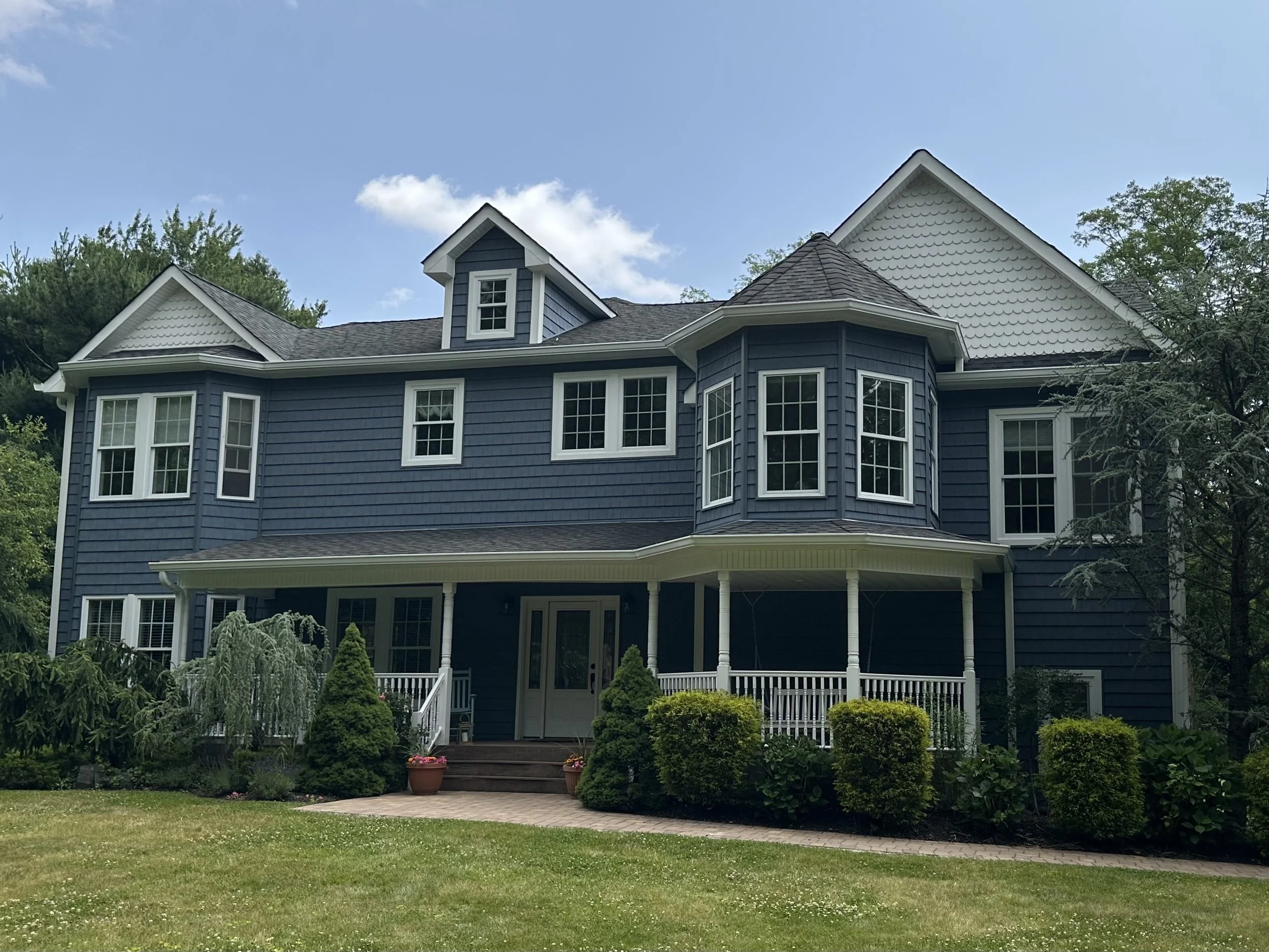A large blue house with multiple gables and white trim, surrounded by a well-maintained lawn and garden with bushes and flowers, under a partly cloudy sky.