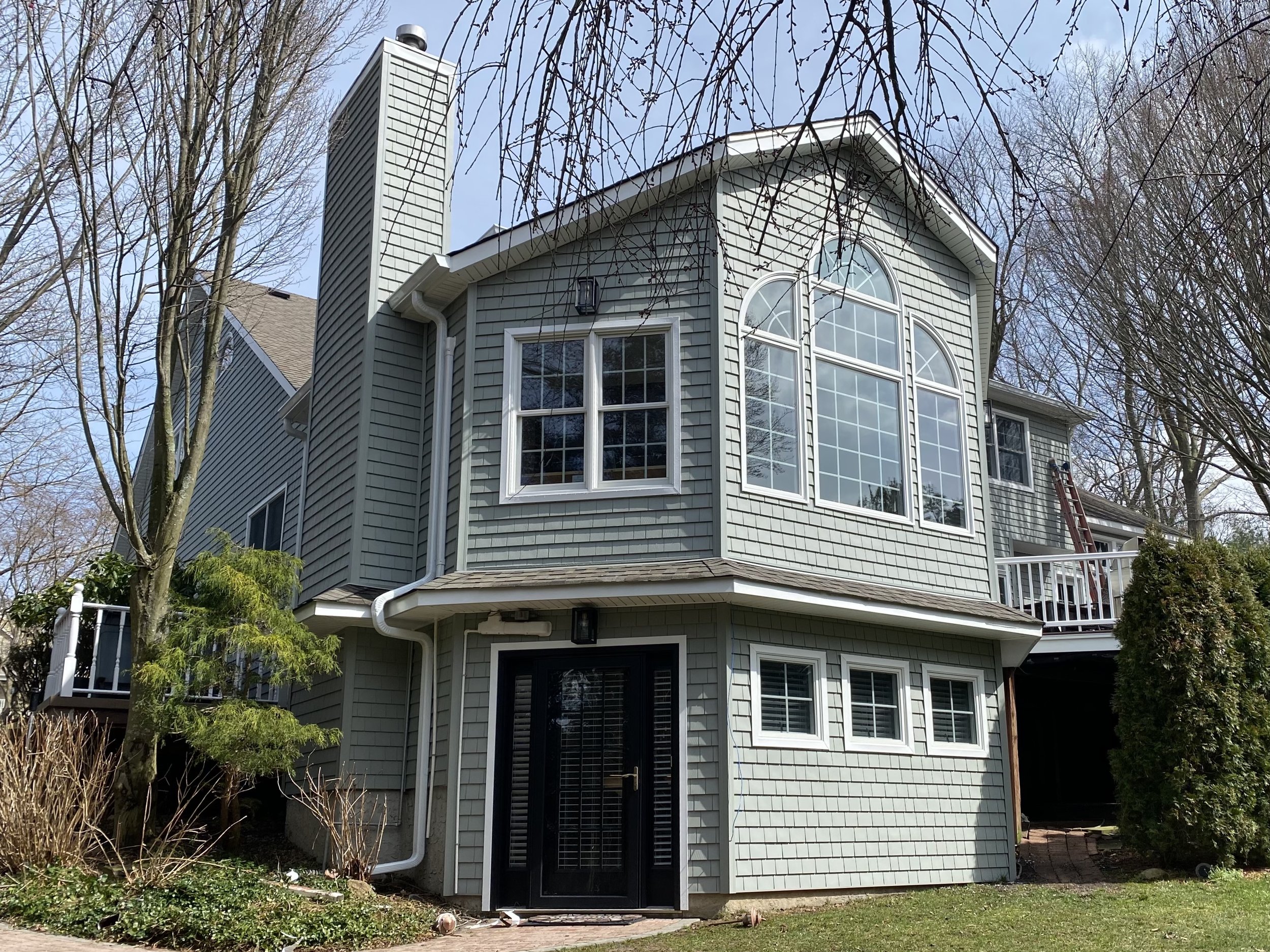 Three-story house with gray siding, large arched windows, and a white gutter system, surrounded by leafless trees and shrubs, with a clear blue sky in the background.