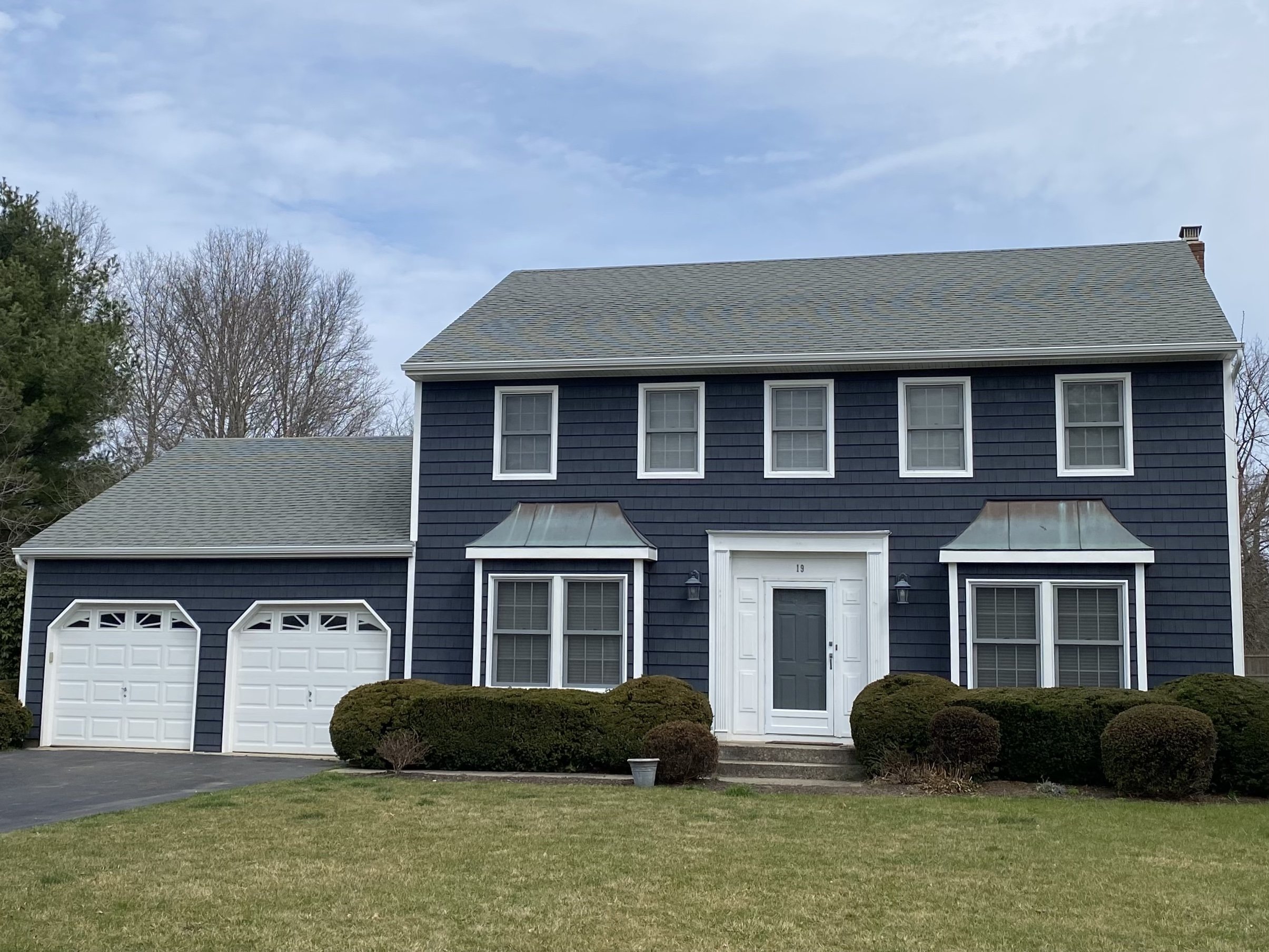 A two-story house with dark blue siding, white trim, and a gray shingle roof. The house features two white garage doors, a front door, and multiple windows. There are small bushes and a lawn in front of the house, with leafless trees in the backgroun