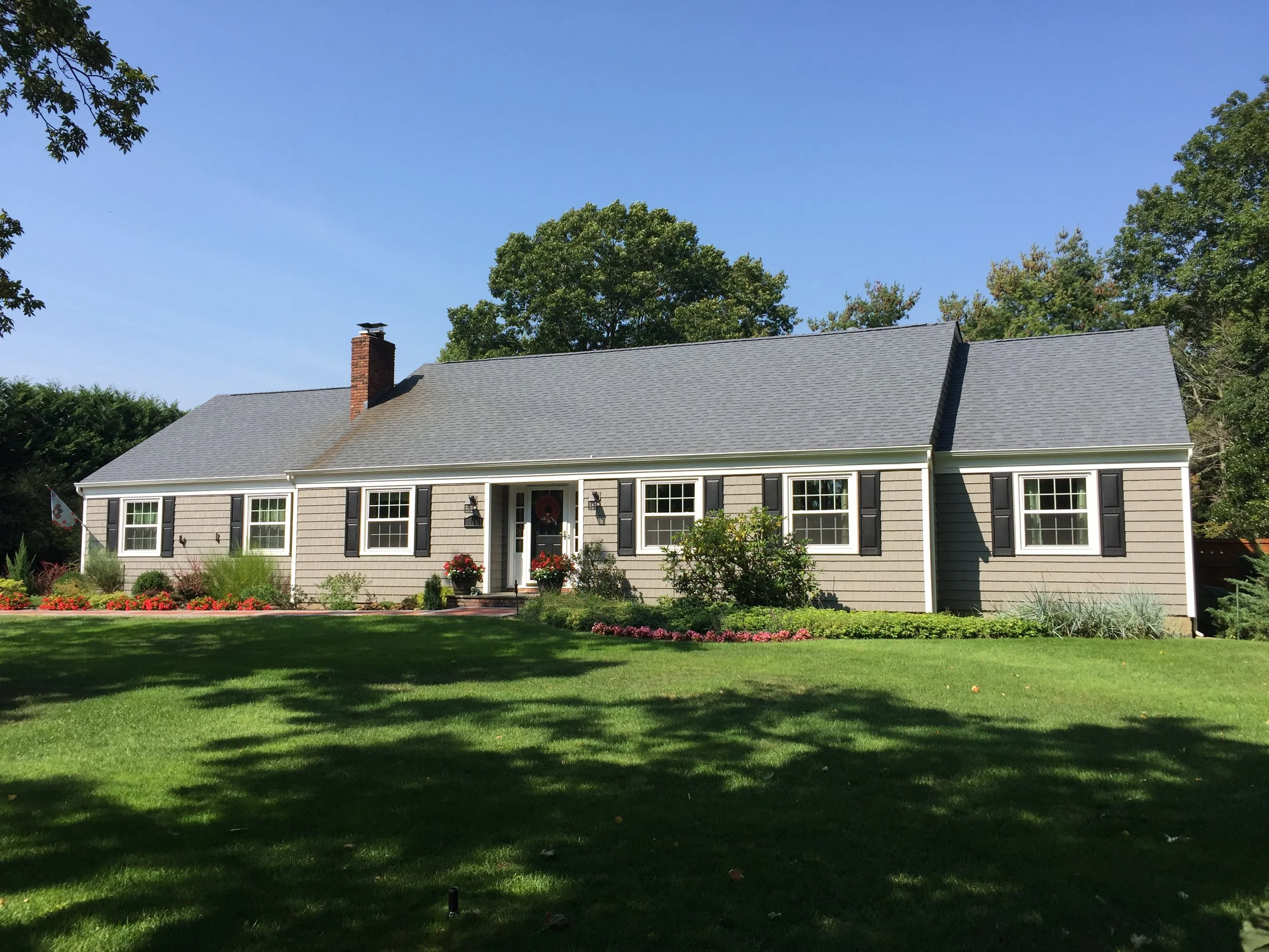 A house with a gray roof, brick chimney, and beige siding, surrounded by a green lawn and trees under a blue sky.