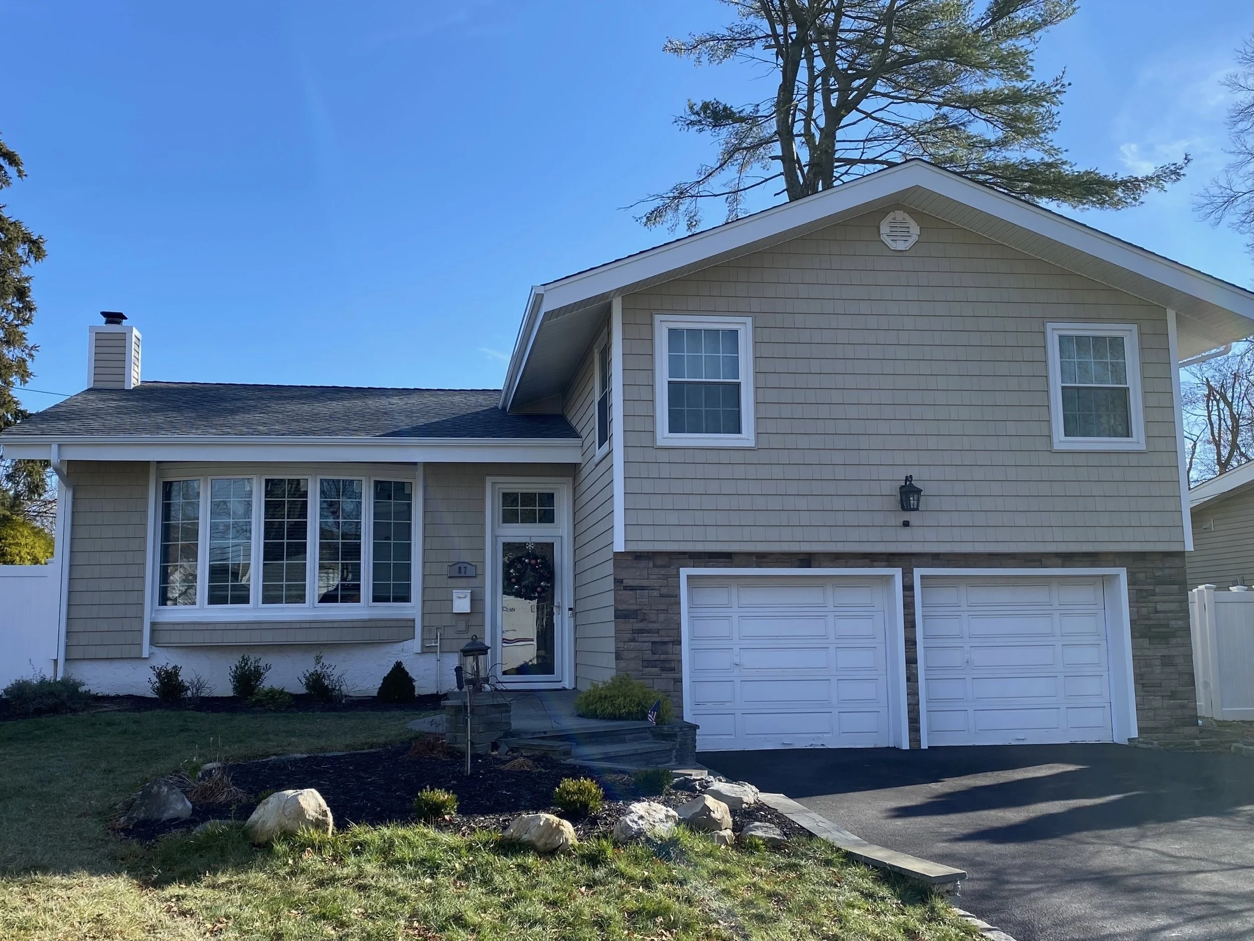 A two-story house with beige siding, white trim, and a gray shingle roof. The house has multiple windows, a front door with a wreath, and a white garage door. There is a small front yard with rocky landscaping, plants, and a black driveway. A large t