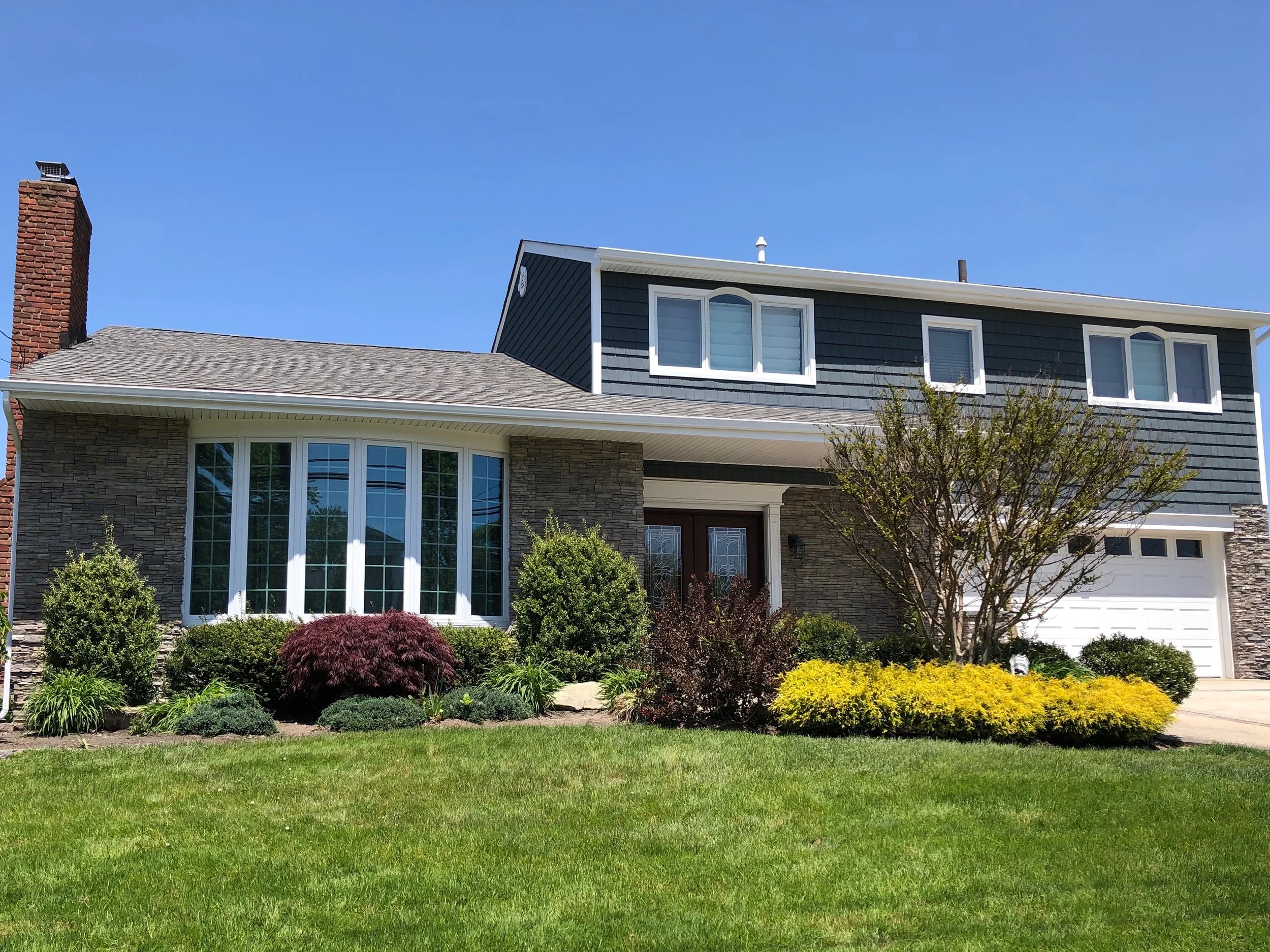 Front view of a two-story house with a brick chimney on the left, large bay window on the left, dark gray siding on the upper level, stone accents on the lower level, and a white garage door on the right. The front yard has well-kept grass, shrubs, a
