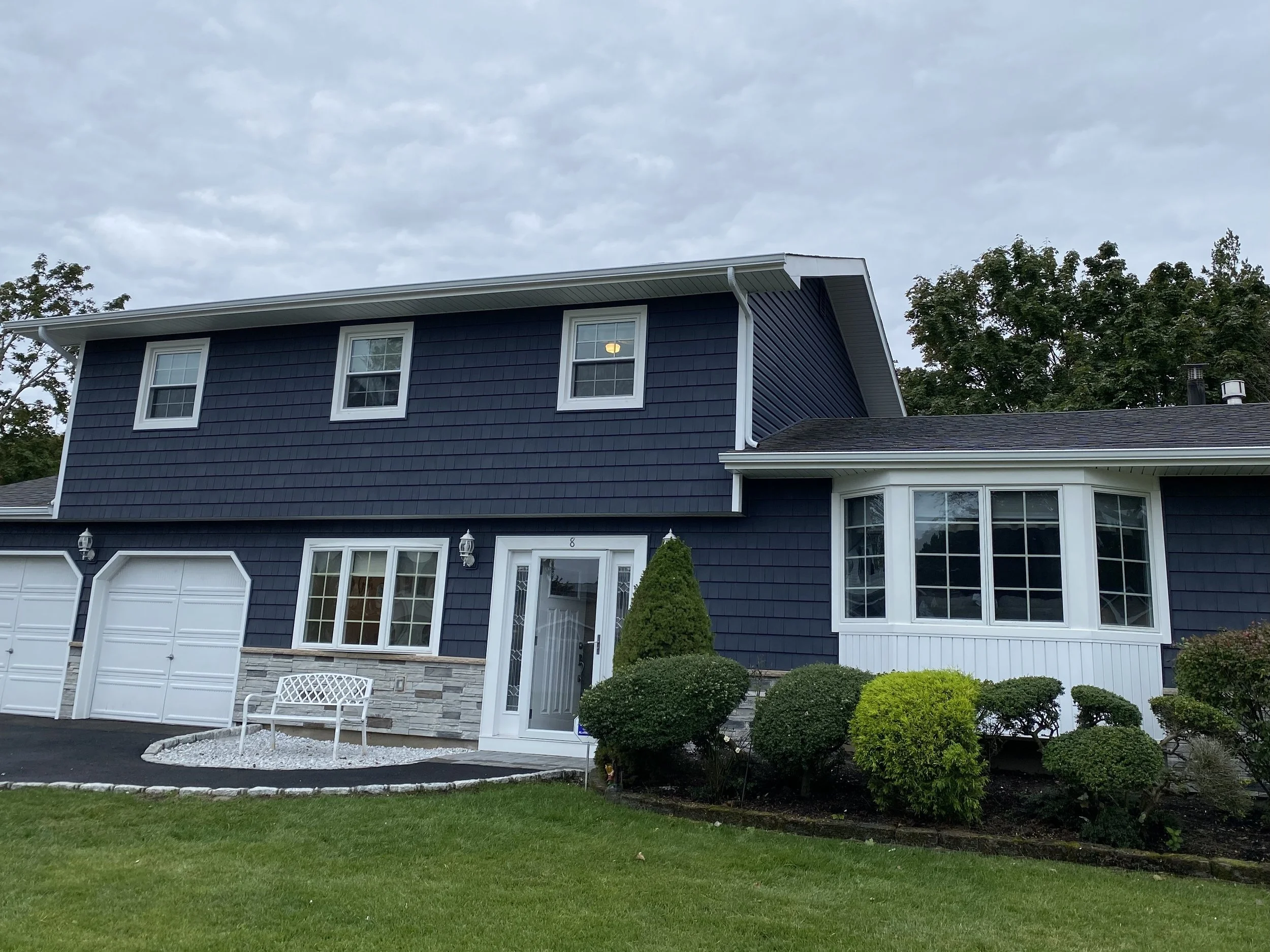 A two-story house with navy blue siding, white trim, and a gray roof. The front yard has a green lawn, decorative bushes, and a white bench near the entrance. The sky is cloudy.