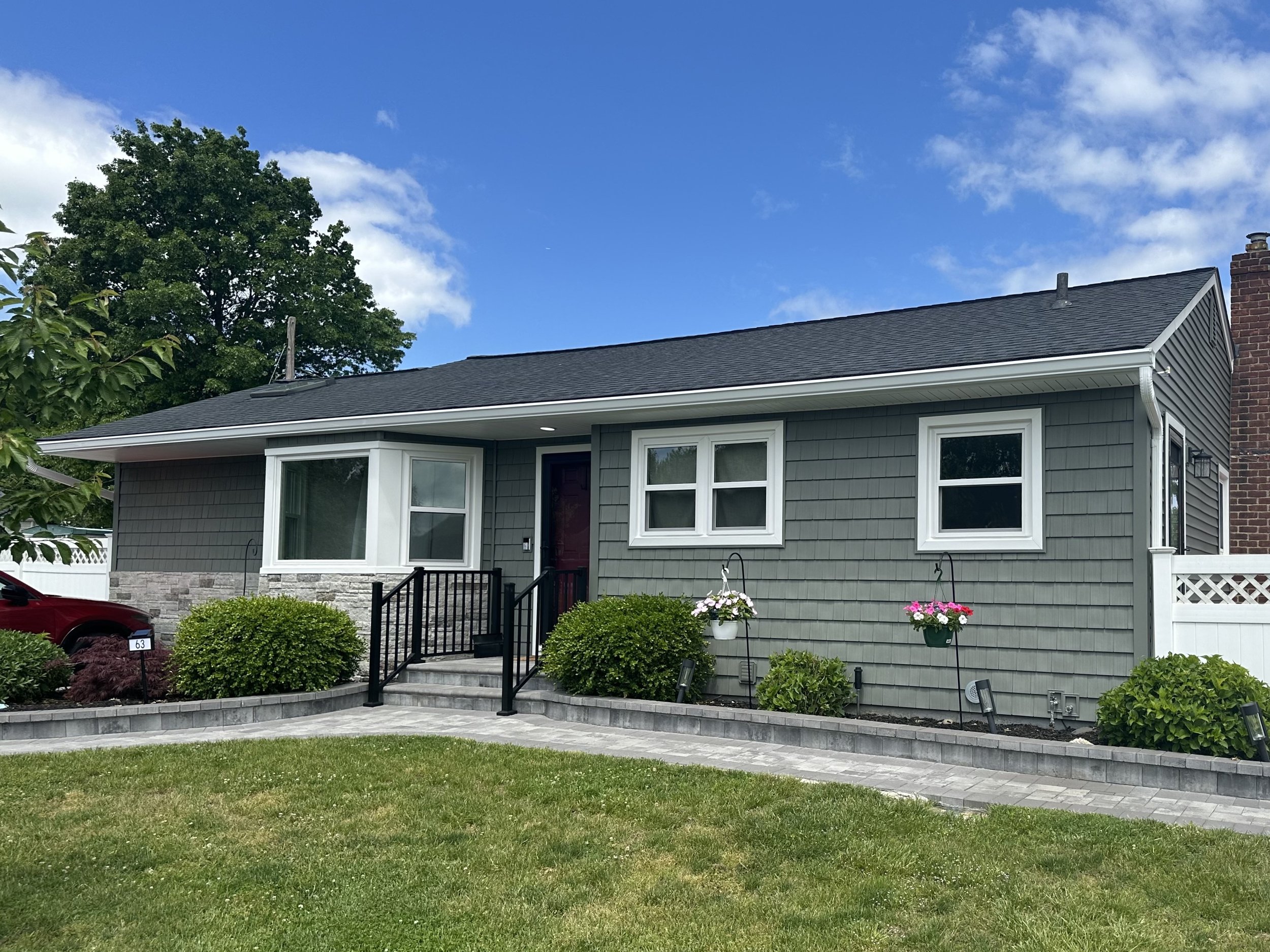 A house with a gray exterior and white window frames, a dark shingled roof, brick chimney, and a front porch with steps, surrounded by well-kept landscaping, including bushes and flowers. There is a blue sky with some clouds and a large green tree in