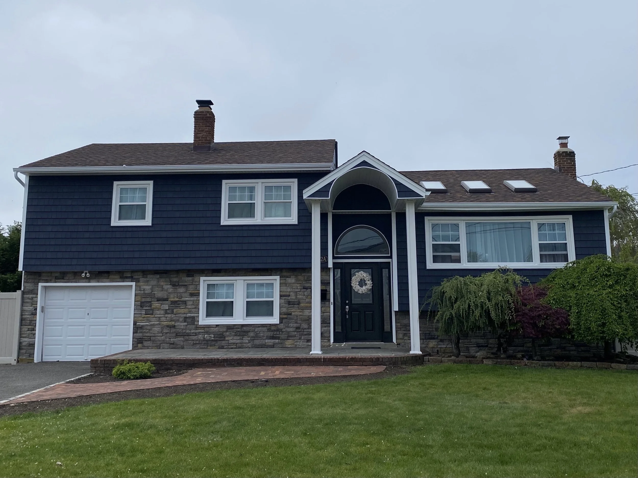 A two-story house with blue siding, stone accents, and a dark garage door, featuring a porch with a black door, multiple windows, and two chimneys on the roof, with a lawn and trees in the foreground under a cloudy sky.