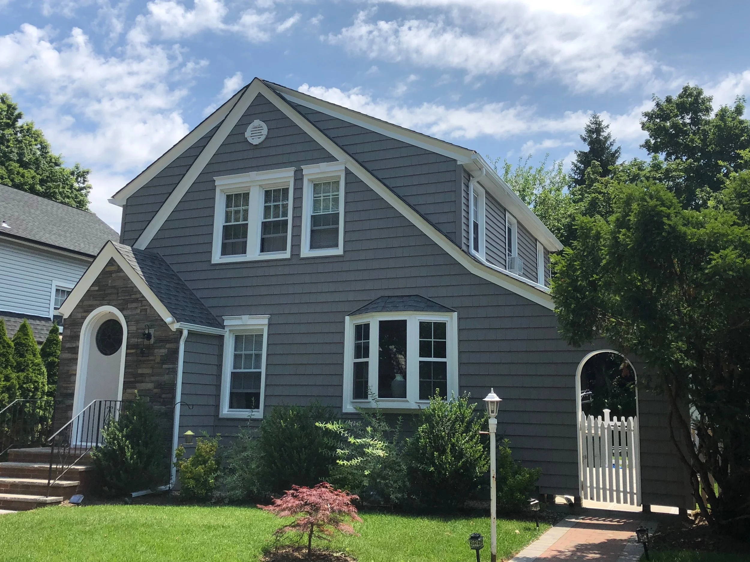 A two-story house with gray siding and white trim, multiple windows, a small front porch with stairs, a white picket fence, and various trees and shrubs in the yard under a partly cloudy sky.