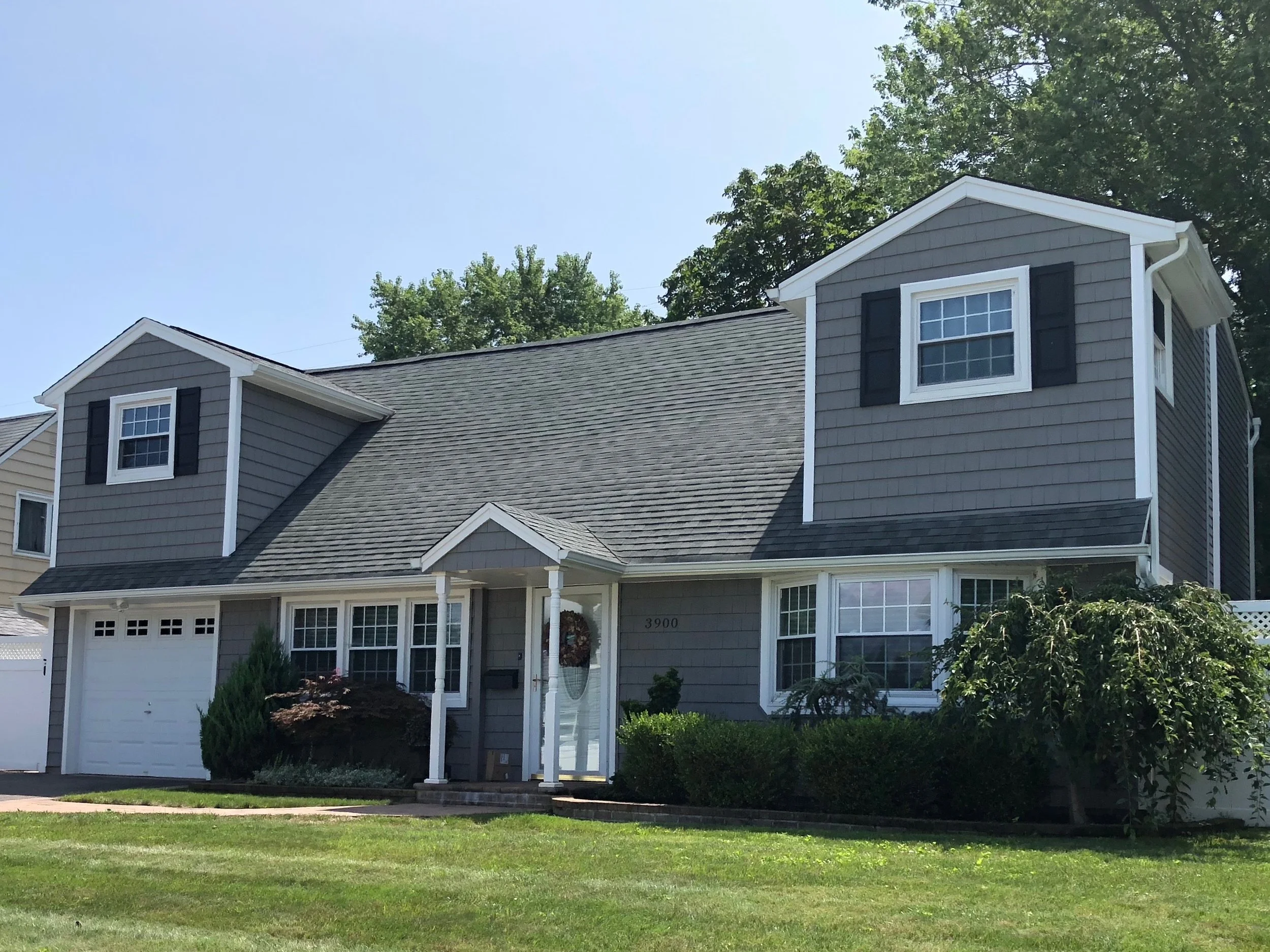 A two-story gray house with a sloped roof and two dormer windows, one with black shutters, a white front porch with columns, and a white garage door, surrounded by green bushes and a lawn, under a clear blue sky.