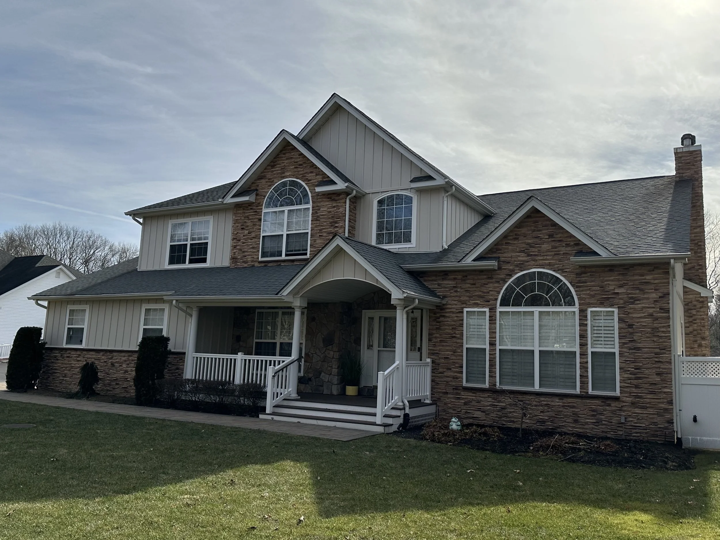 A two-story house with a combination of siding and brick exterior, multiple windows, a front porch with white railings, and a sloped roof with a chimney. The house is surrounded by a well-manicured lawn with bushes.