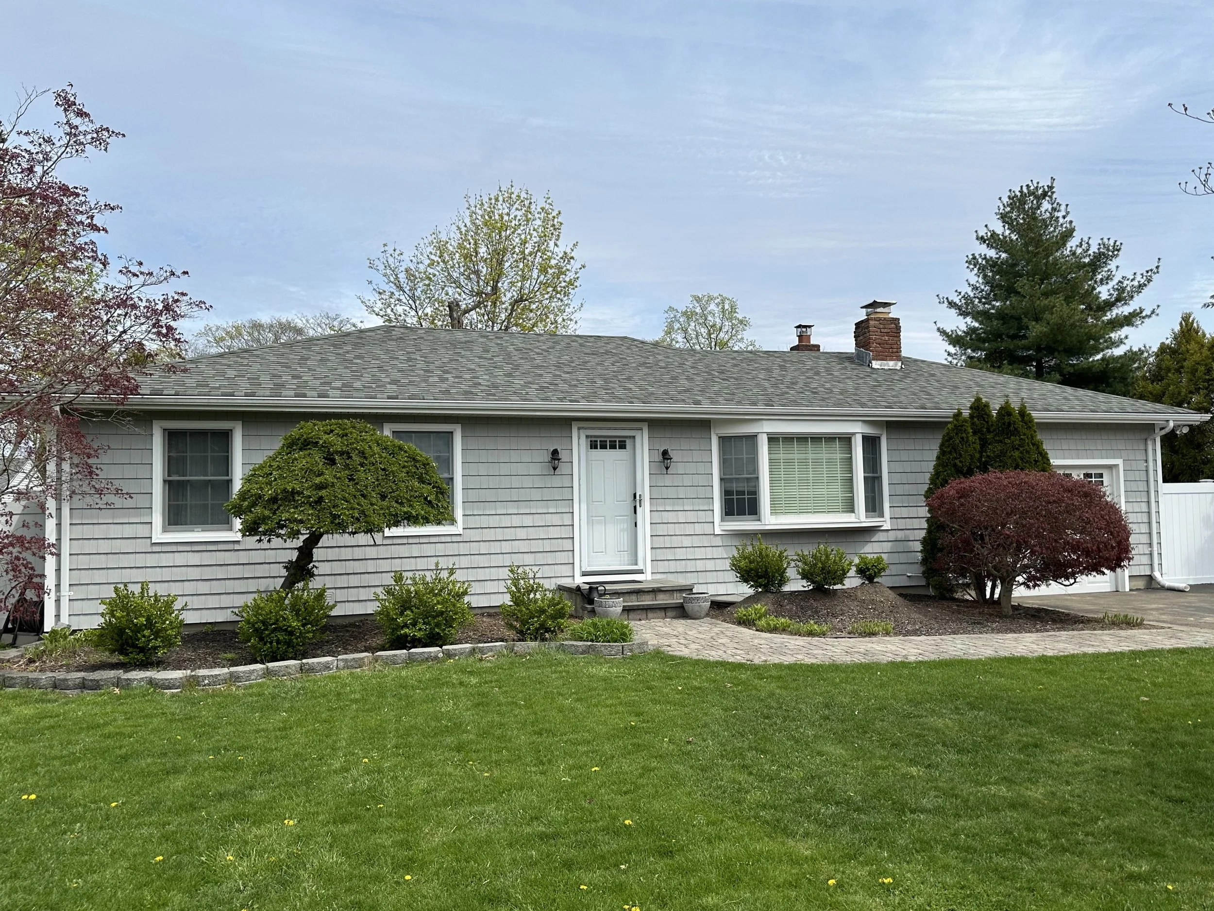 A house with a gray shingled roof and white siding, surrounded by green grass, bushes, and trees, under a partly cloudy sky.