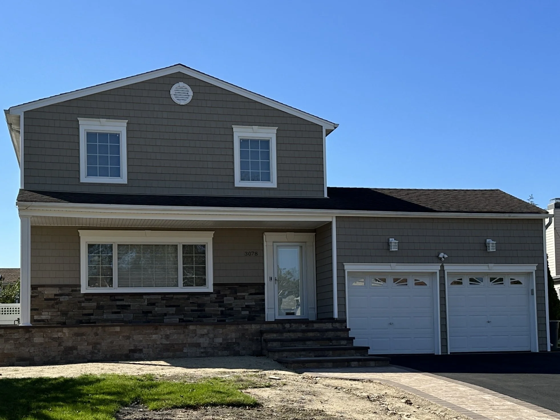 A two-story house with gray siding, white window frames, and a garage door, with a small lawn and stone steps leading to the front door, under a clear blue sky.