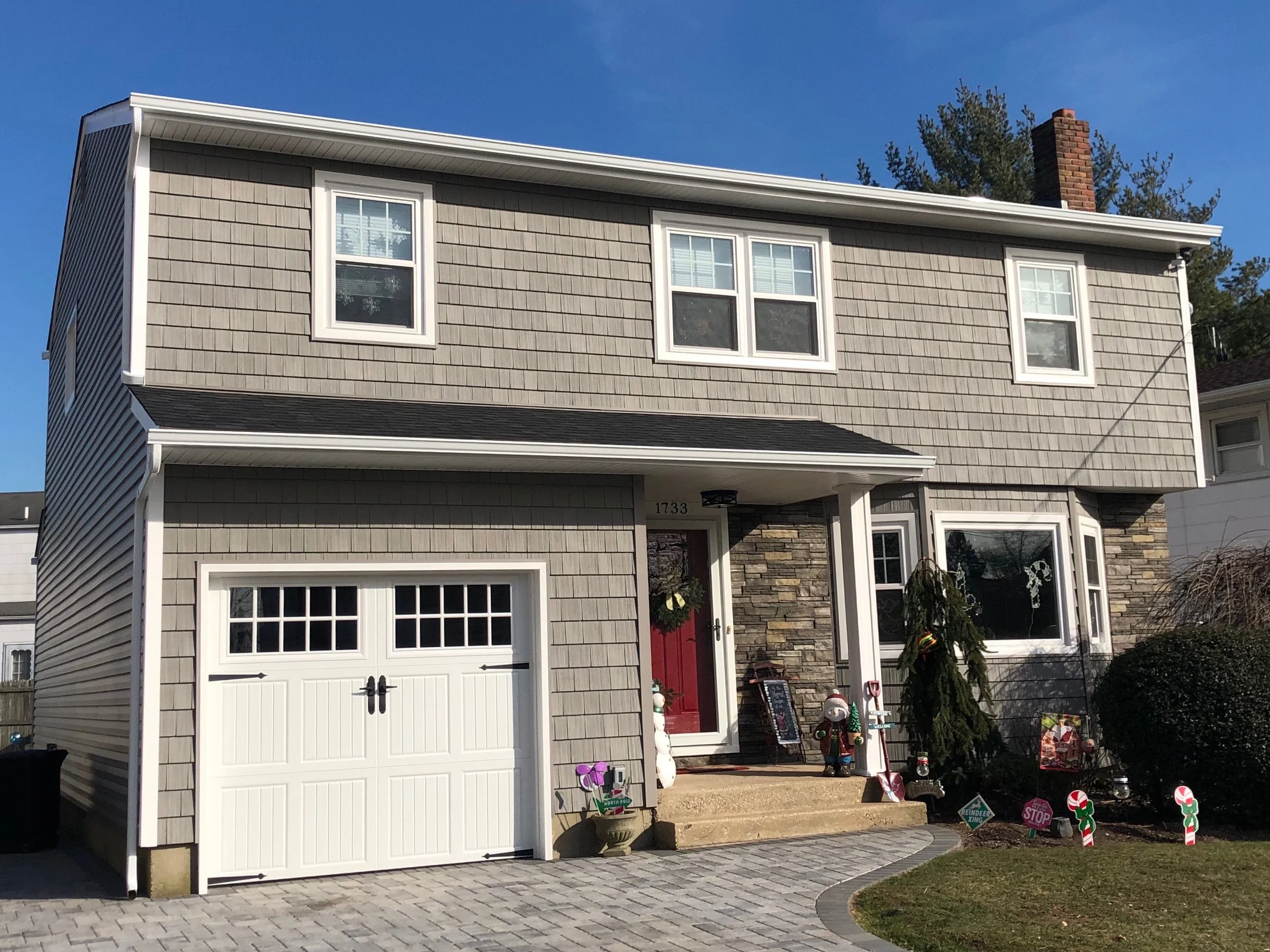 Front view of a two-story house with gray shingle siding, white trim, and a red front door. Christmas decorations and holiday signs are displayed outside. There is a garage with a white door, and a brick chimney on the roof. The lawn is decorated wit