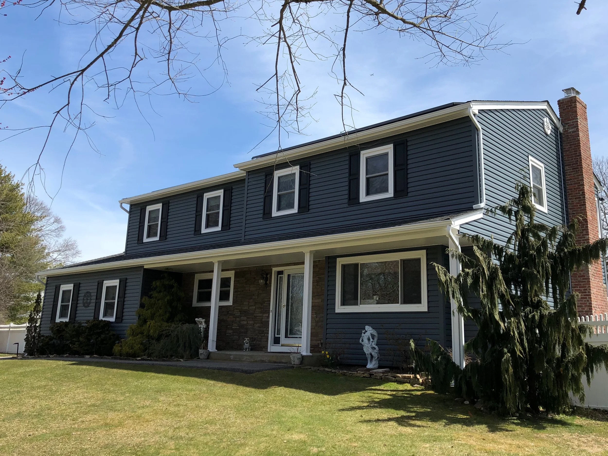 A two-story house with blue siding, black shutters, and white trim, featuring a brick chimney on the right side. The front yard has a well-maintained lawn with some bushes and statues, and a porch with white support columns. Bare tree branches extend