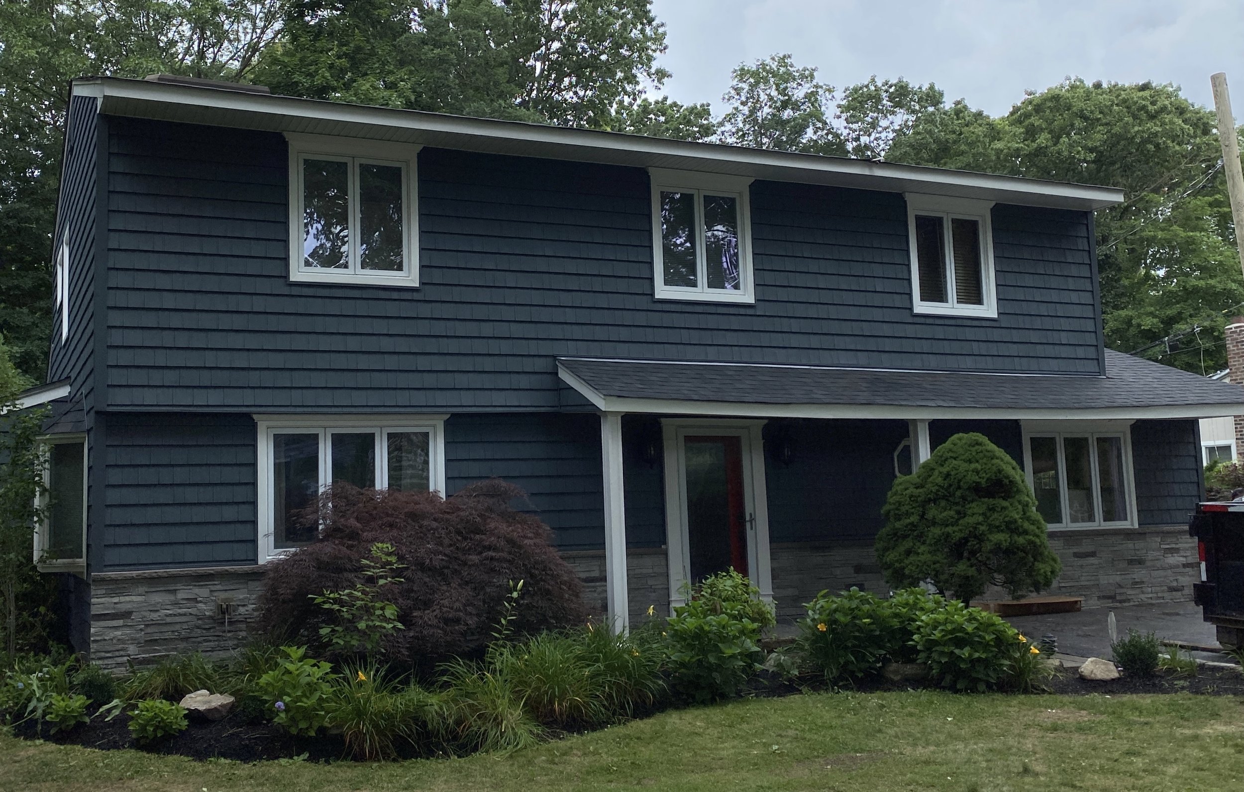 A two-story house with dark blue siding, multiple white-framed windows, and a front porch. The yard has green grass, bushes, and small trees, with a backdrop of tall trees and power lines.
